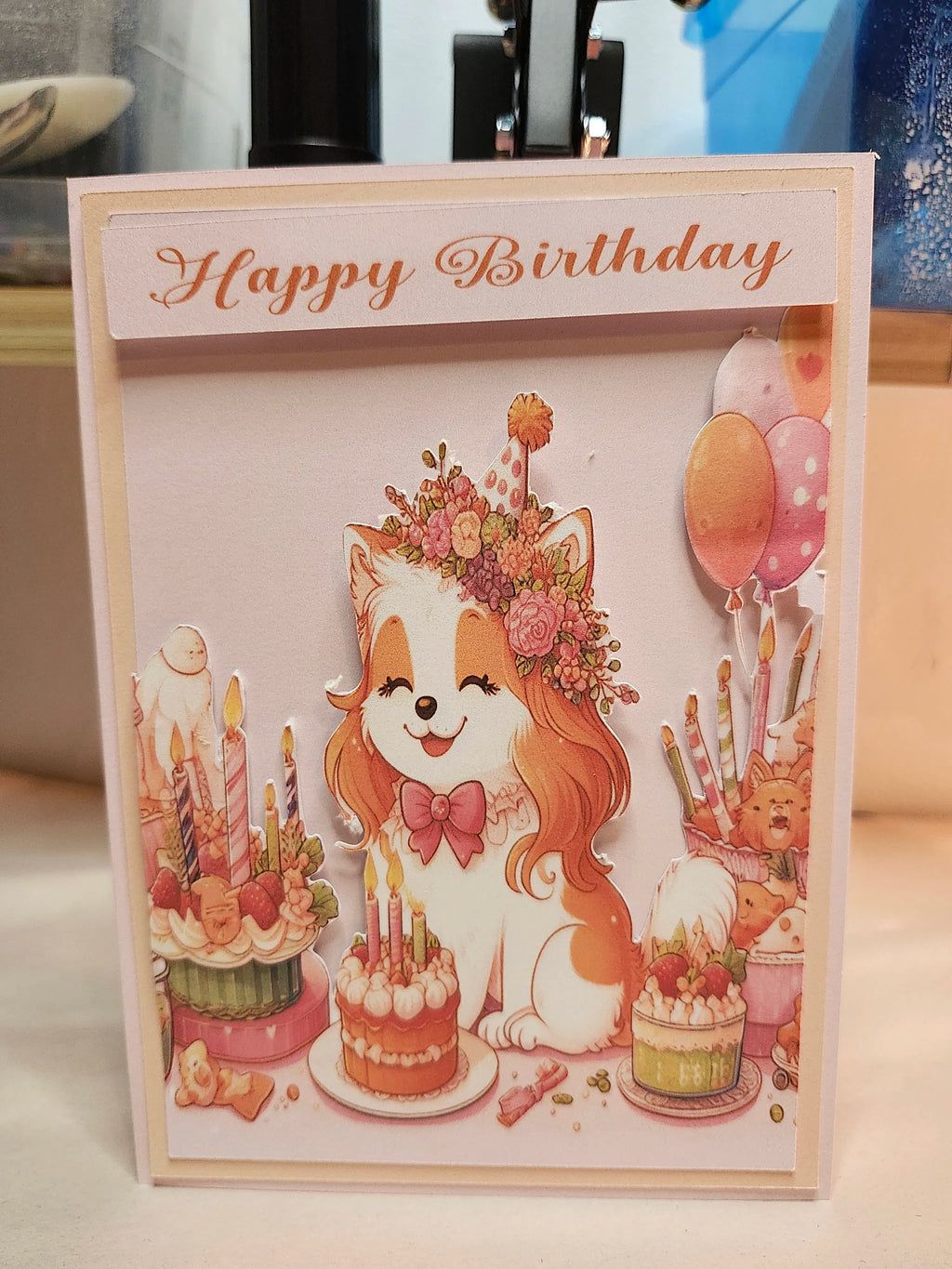 A handmade 'Happy Birthday' card featuring a cute cartoon dog wearing a floral crown and bow tie, surrounded by birthday cakes, candles, balloons, and a teddy bear. The card is photographed standing on a light-colored surface with a blurred background.