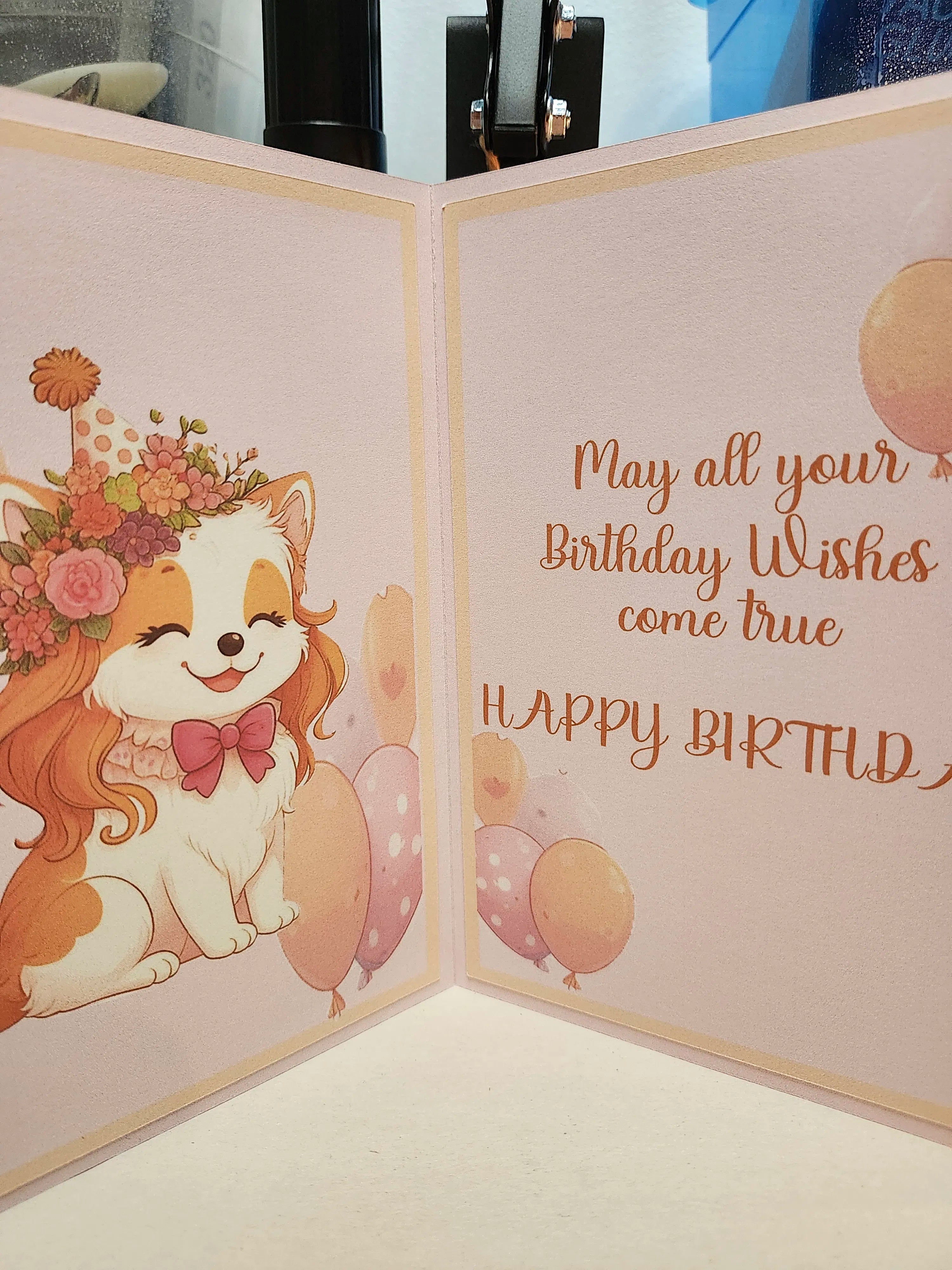 A handmade paper birthday card shown open, featuring a cute cartoon dog wearing a floral hat and bow tie on the left panel, and the text 'May all your Birthday Wishes come true HAPPY BIRTHDAY' on the right panel.