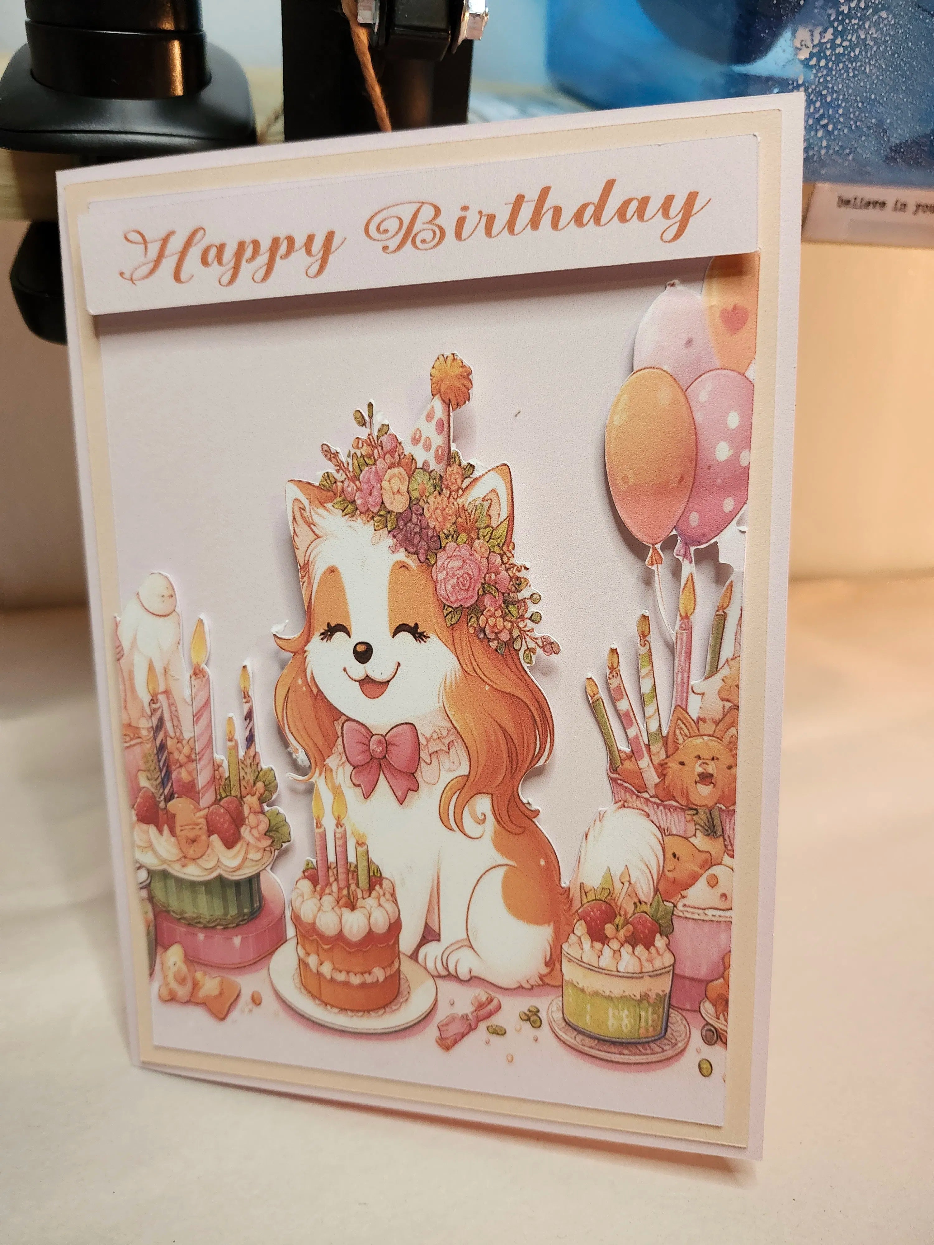 A handmade 'Happy Birthday' greeting card featuring a cute cartoon fox wearing a floral crown and bow tie, surrounded by birthday cakes, candles, balloons, and teddy bears. The card is photographed standing on a light-colored surface with some background clutter visible.