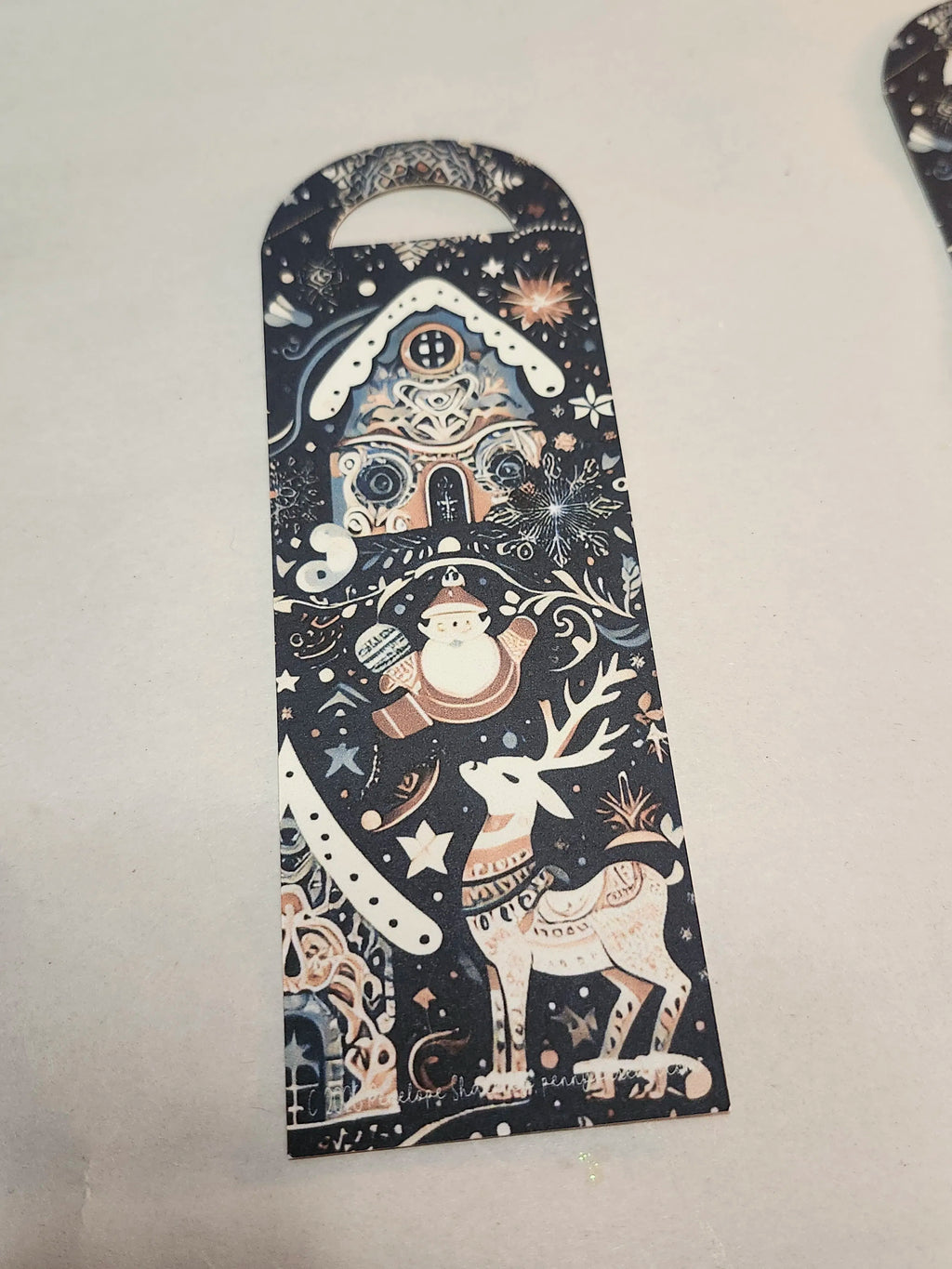 A long, narrow Christmas-themed card or tag with a rounded top handle, featuring a dark navy background decorated with a whimsical winter scene including a Santa Claus, reindeer, snowflake, and gingerbread house in cream, white, and muted pink tones.