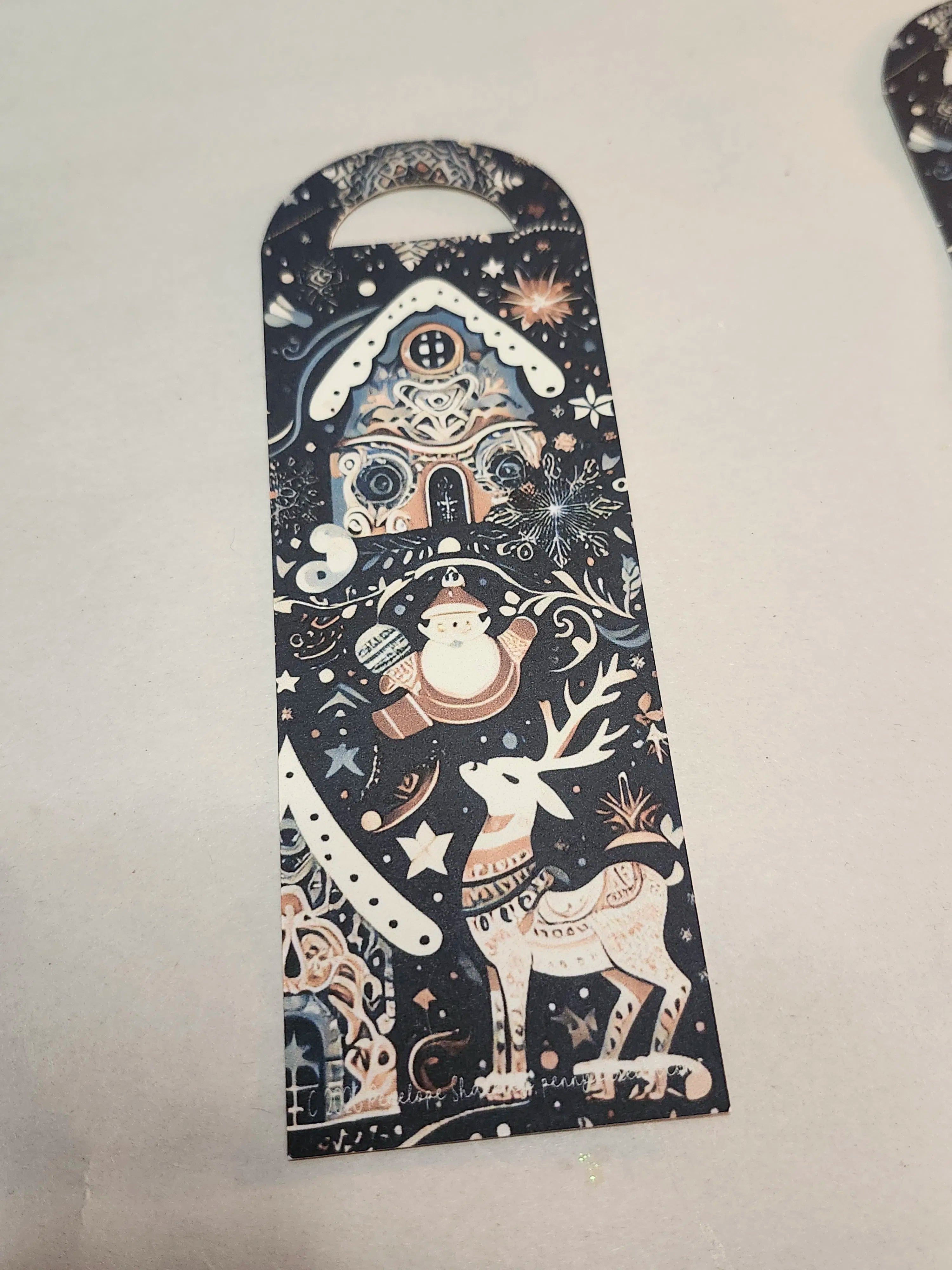 A long, narrow Christmas-themed card or tag with a rounded top handle, featuring a dark navy background decorated with a whimsical winter scene including a Santa Claus, reindeer, snowflake, and gingerbread house in cream, white, and muted pink tones.