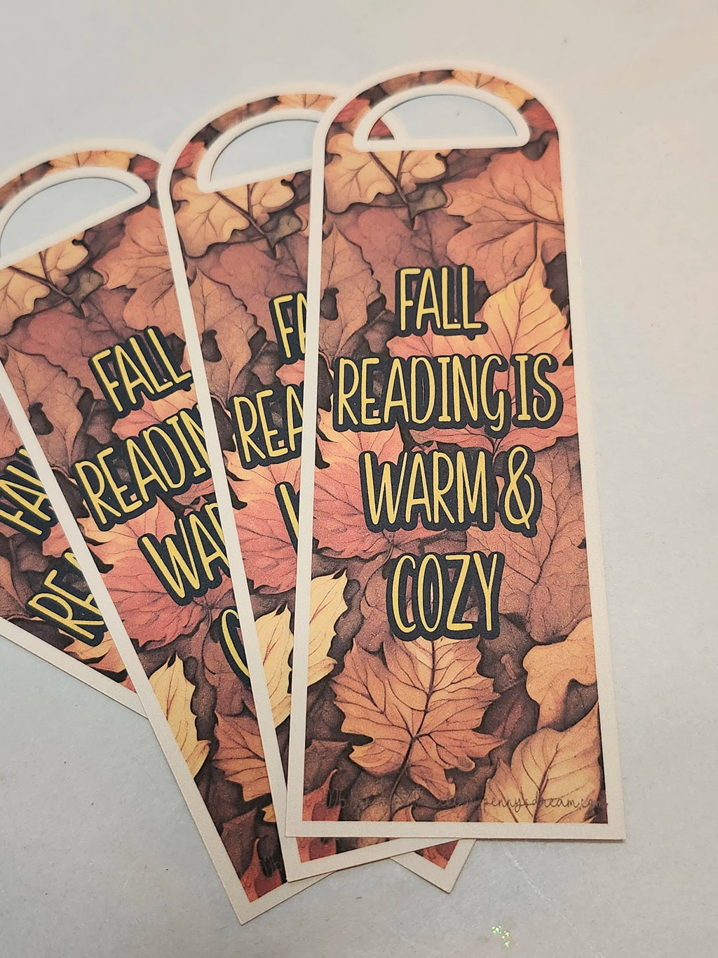 Three arched bookmarks with a fall leaf pattern, fanned out on a light gray surface. The bookmarks feature the text 'FALL READING IS WARM & COZY' in a yellow outlined font.