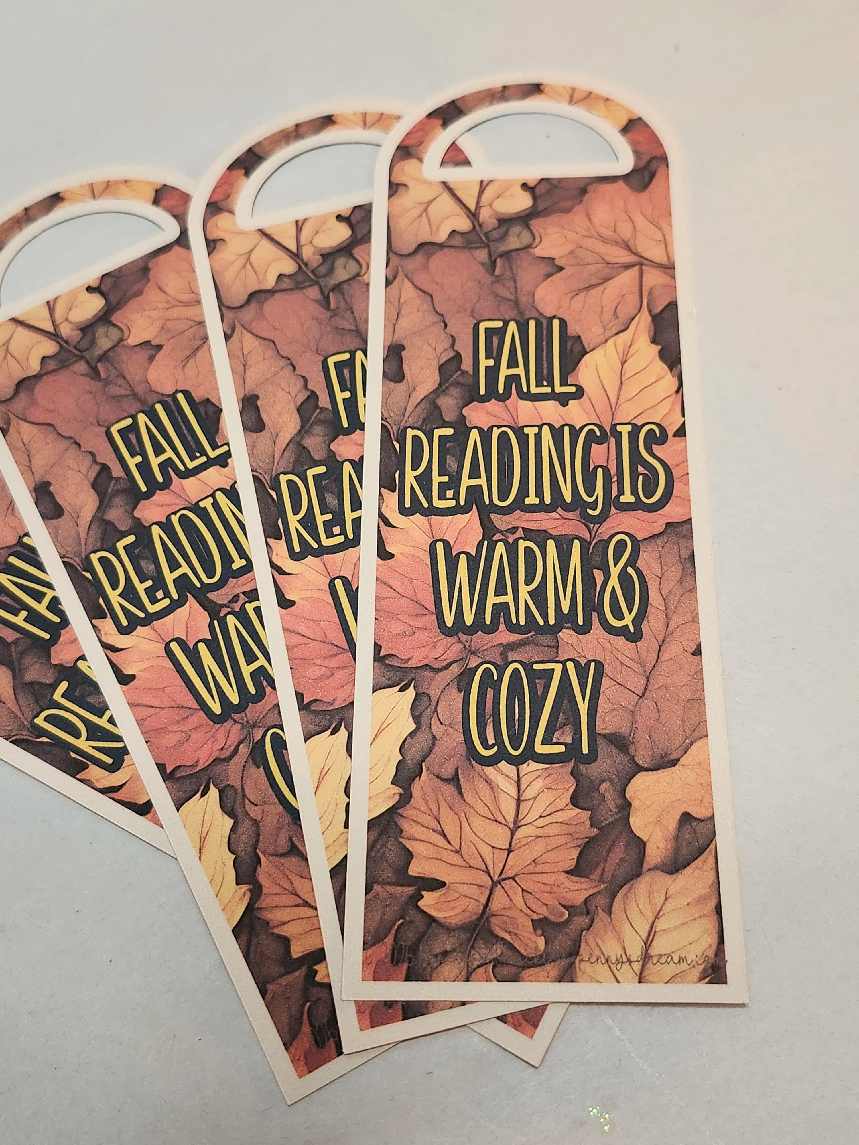 Three arched bookmarks with a fall leaf pattern, fanned out on a light gray surface. The bookmarks feature the text 'FALL READING IS WARM & COZY' in a yellow outlined font.
