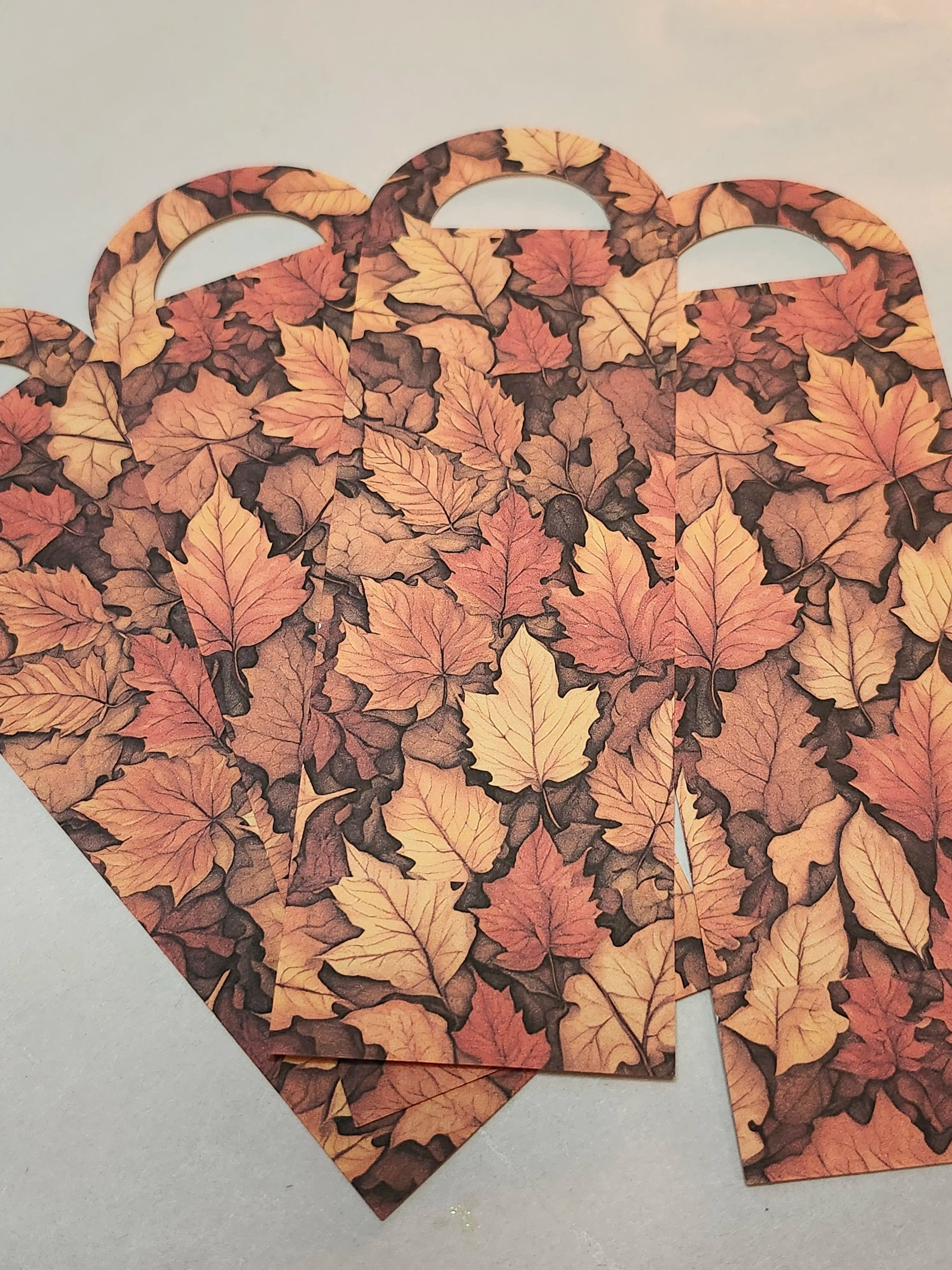 Three paper gift bags with handles, printed with an autumn leaf pattern in shades of orange, brown, and beige, arranged on a light gray surface.