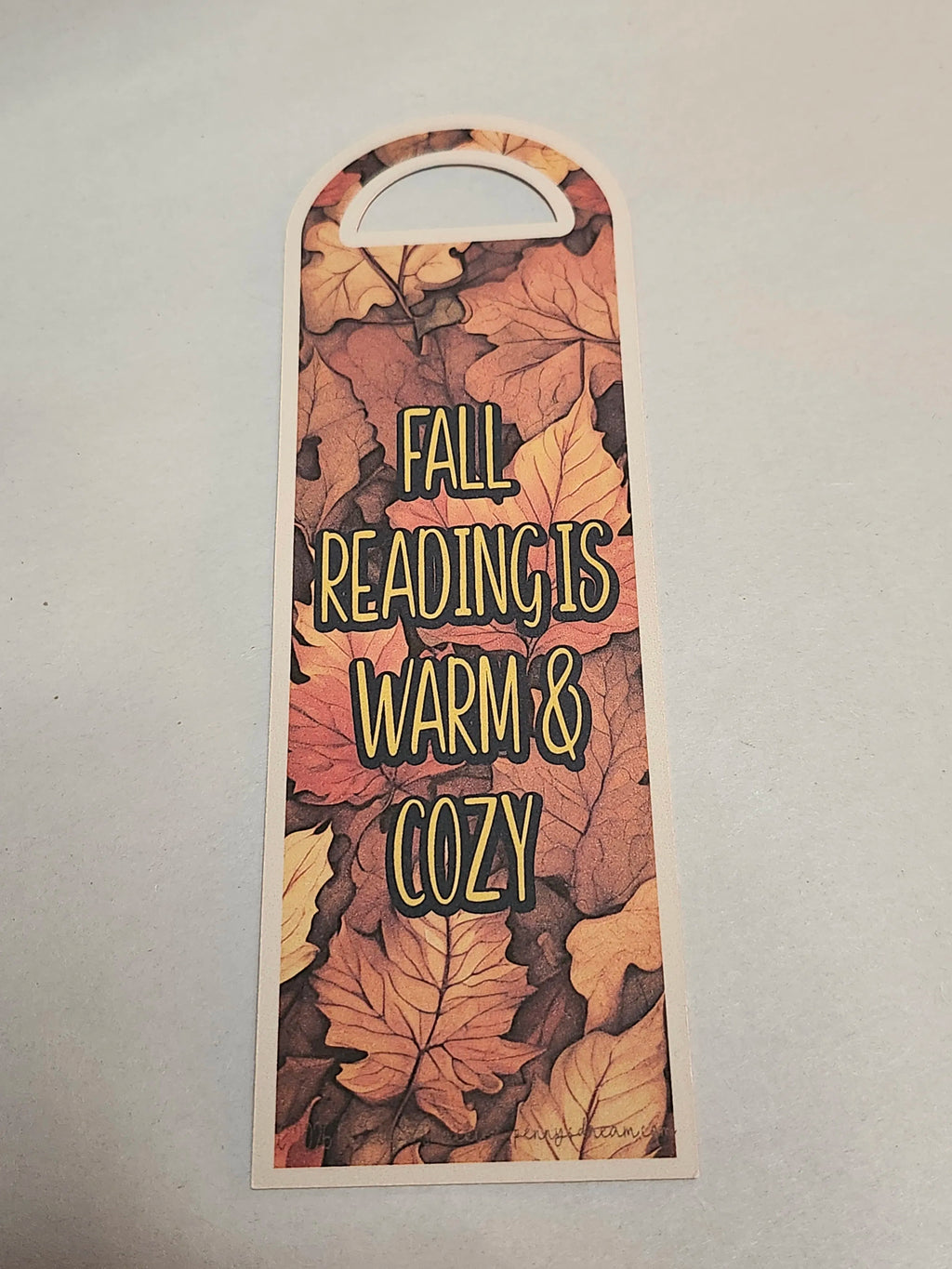 A tall, narrow decorative tag or bookmark with an arched top and a die-cut handle, printed with overlapping autumn leaves and the phrase “FALL READING IS WARM & COZY” in yellow outlined lettering, shown on a light gray background.