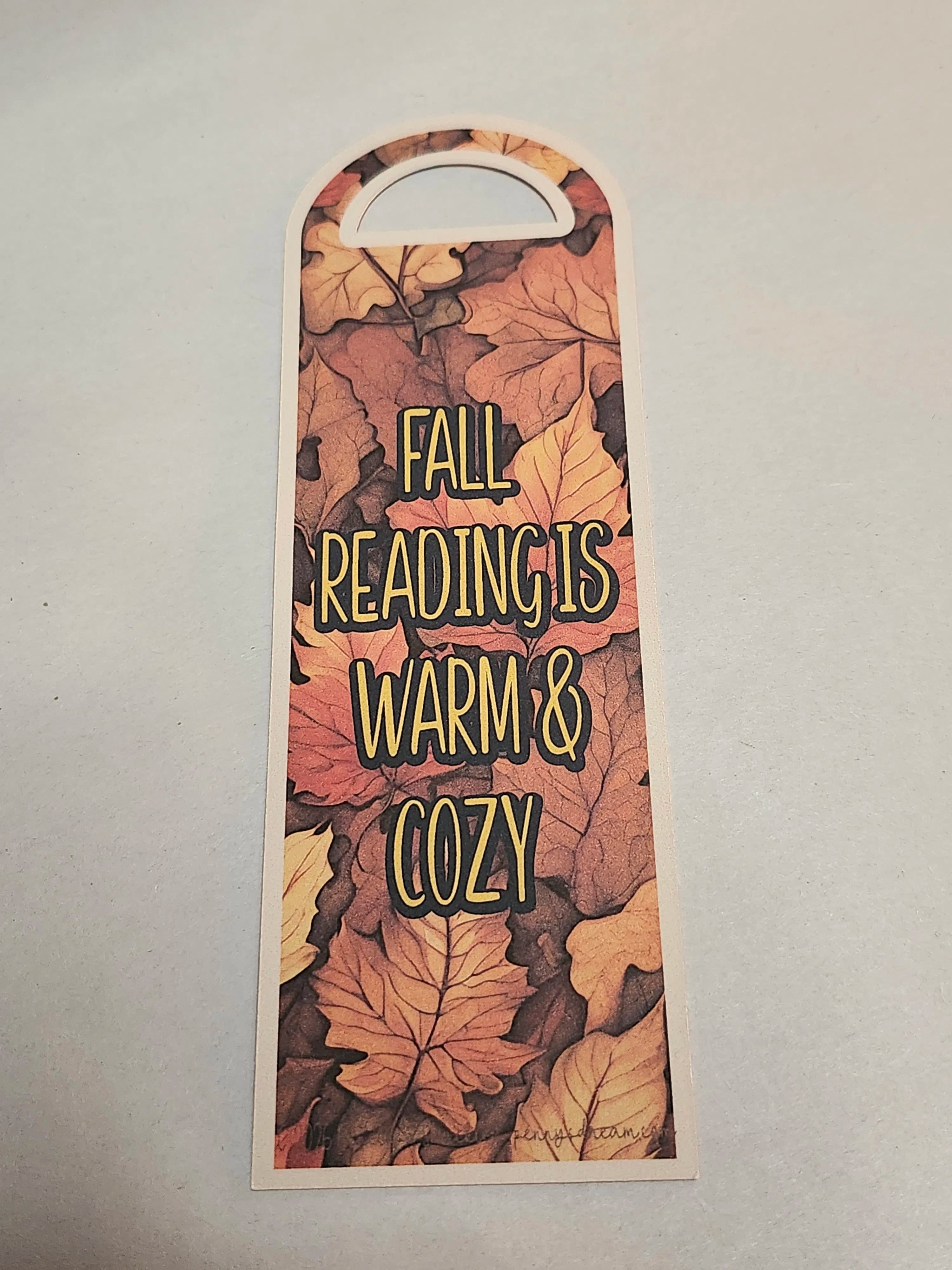 A tall, narrow decorative tag or bookmark with an arched top and a die-cut handle, printed with overlapping autumn leaves and the phrase “FALL READING IS WARM & COZY” in yellow outlined lettering, shown on a light gray background.