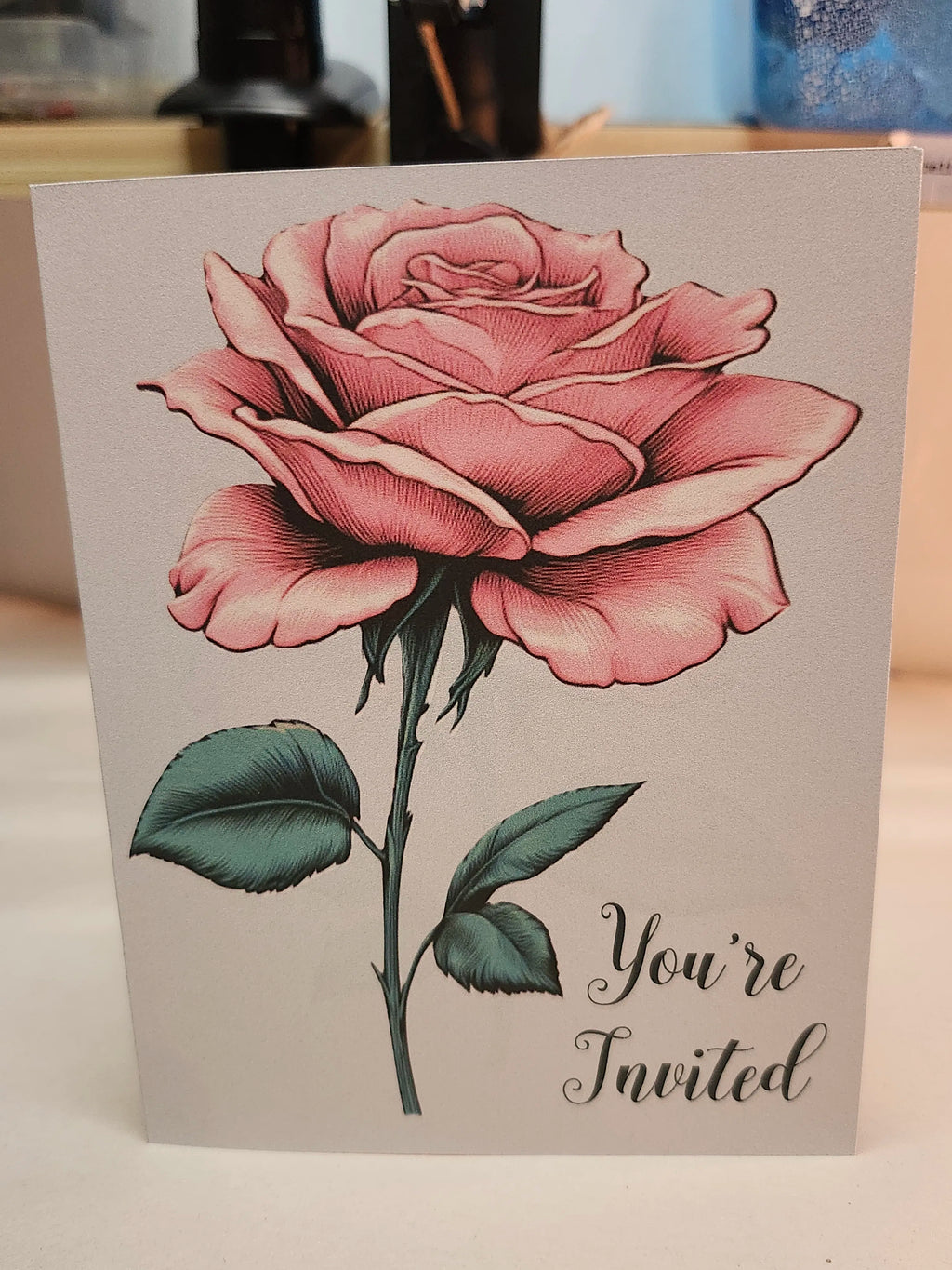 A square greeting card featuring a detailed pink rose illustration with green leaves and the text 'You're Invited' in black script, photographed standing upright on a light surface.