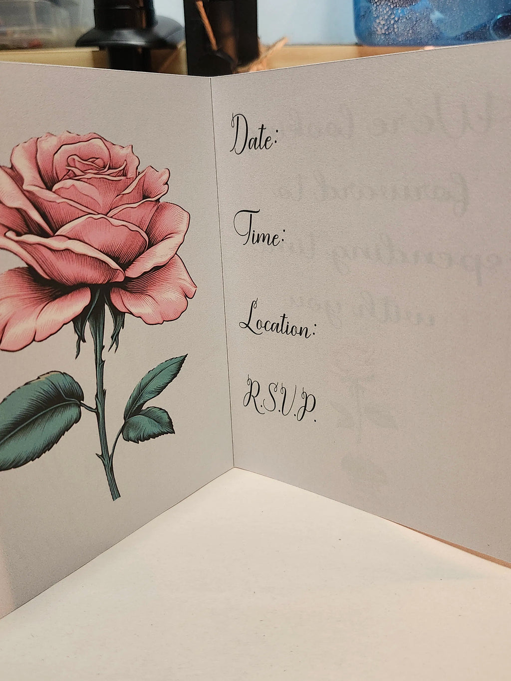 An open invitation card featuring a detailed pink rose illustration on the left panel and printed event details like Date, Time, Location, and RSVP on the right panel.