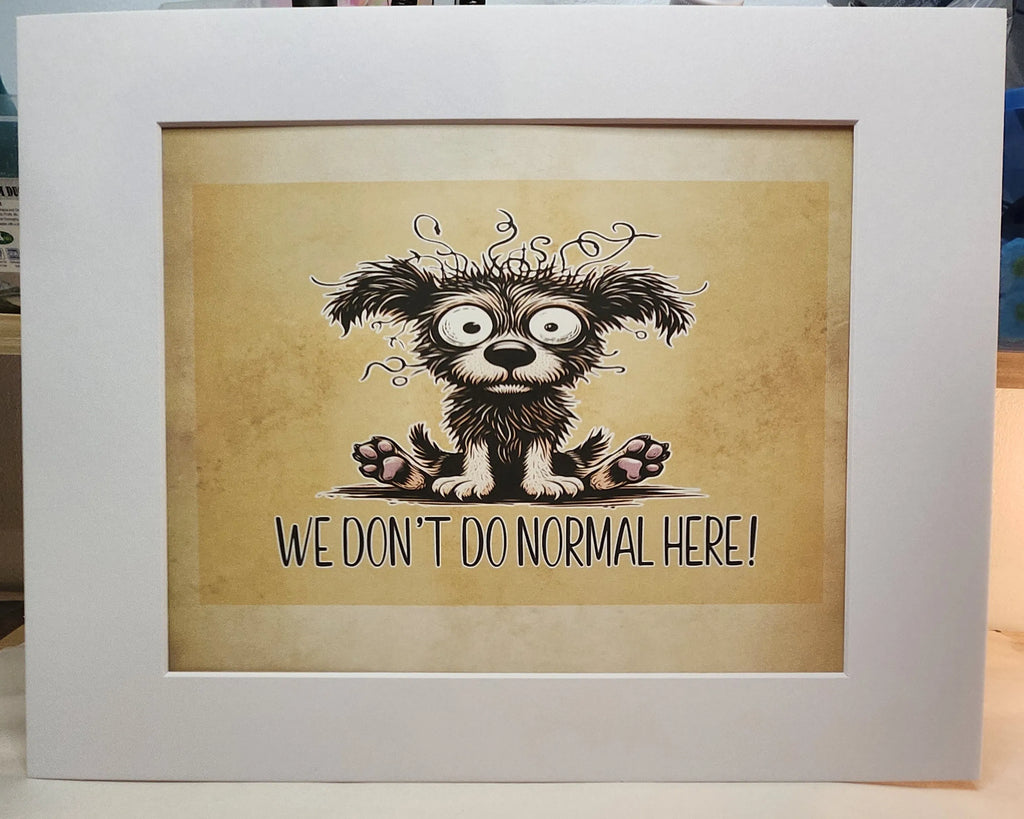 A framed art print featuring a cartoon illustration of a fluffy black and white dog with wide eyes and the text 'WE DON'T DO NORMAL HERE!' on a beige background, enclosed in a white frame.