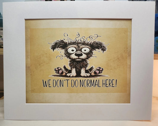 A framed art print featuring a cartoon illustration of a fluffy black and white dog with wide eyes and the text 'WE DON'T DO NORMAL HERE!' on a beige background, enclosed in a white frame.