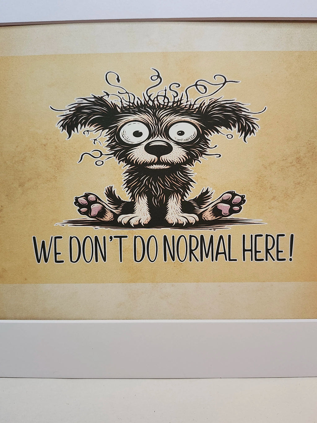 A framed art print featuring a cartoon illustration of a shaggy black and white dog with wild hair and the text 'WE DON'T DO NORMAL HERE!' on a textured beige background.