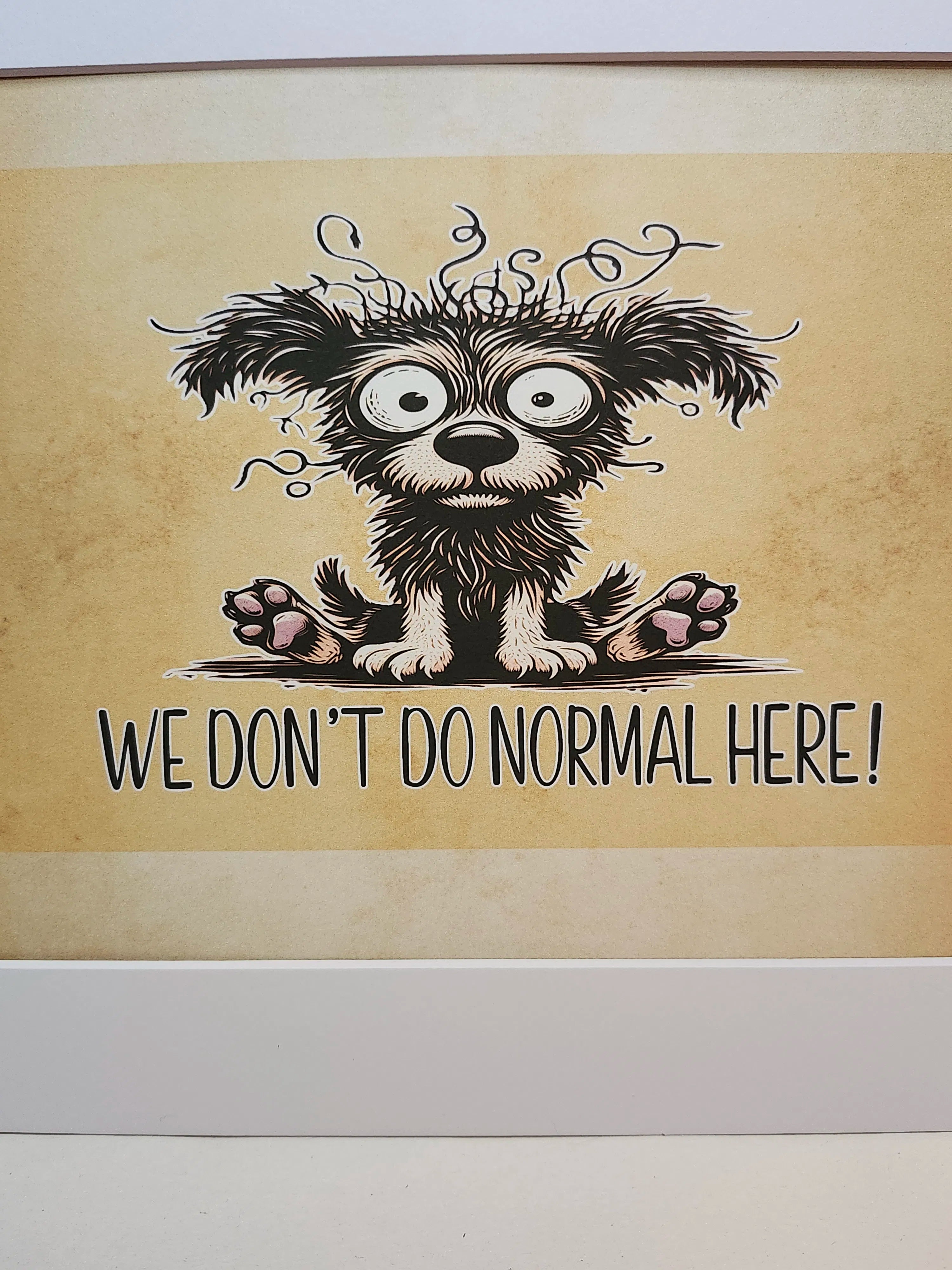 A framed art print featuring a cartoon illustration of a shaggy black and white dog with wild hair and the text 'WE DON'T DO NORMAL HERE!' on a textured beige background.