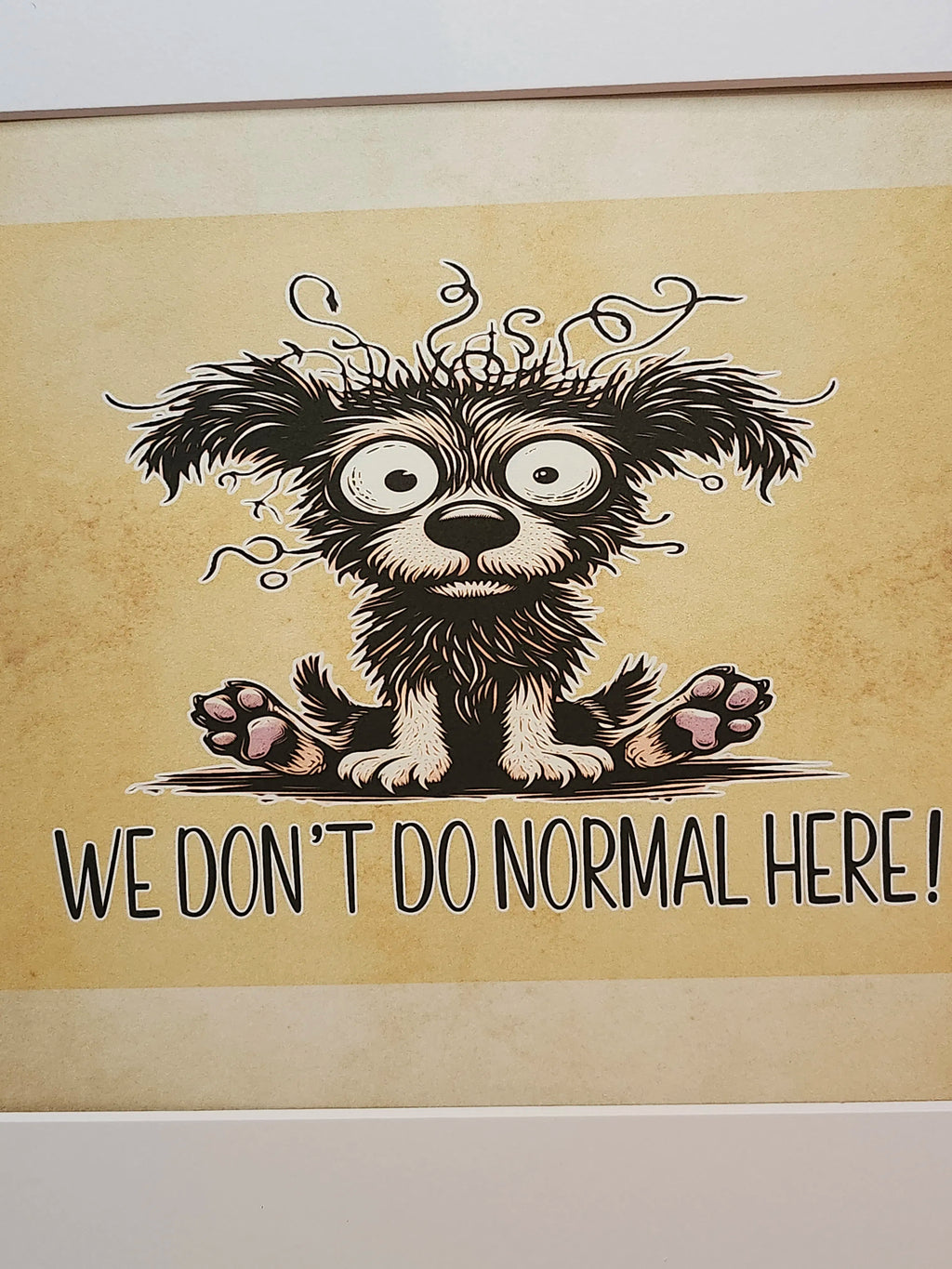 A framed art print featuring a cartoon illustration of a shaggy black and white dog with messy hair and the text 'WE DON'T DO NORMAL HERE!' on a beige background.