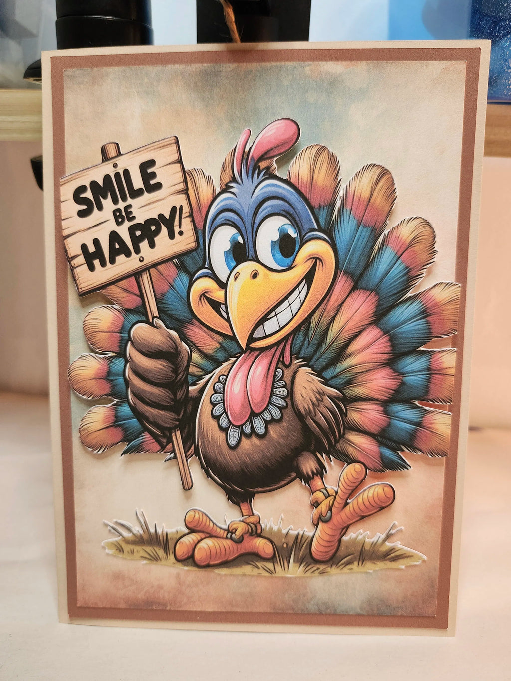 A square greeting card featuring a cartoon turkey holding a sign that reads 'SMILE BE HAPPY!' against a neutral indoor background.