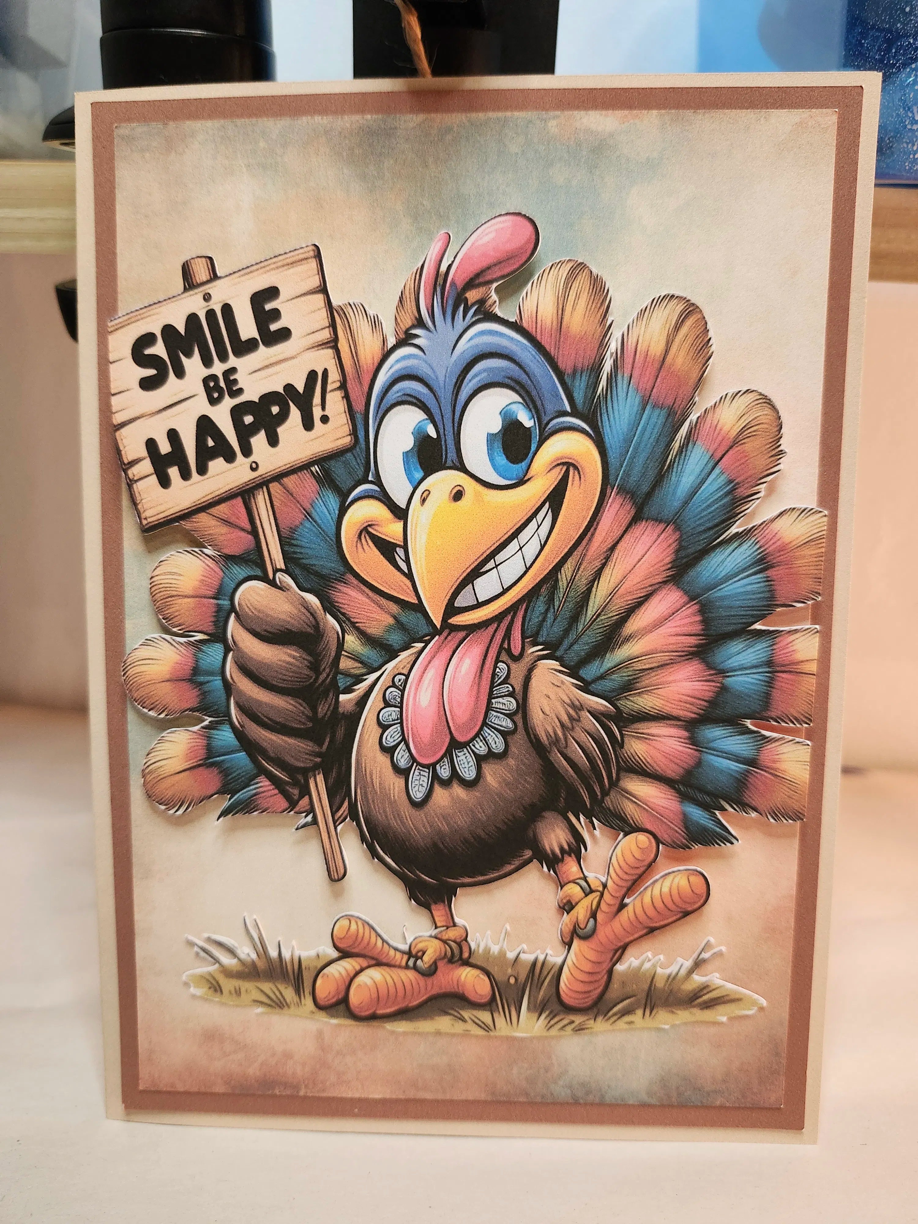 A square greeting card featuring a cartoon turkey holding a sign that reads 'SMILE BE HAPPY!' against a neutral indoor background.