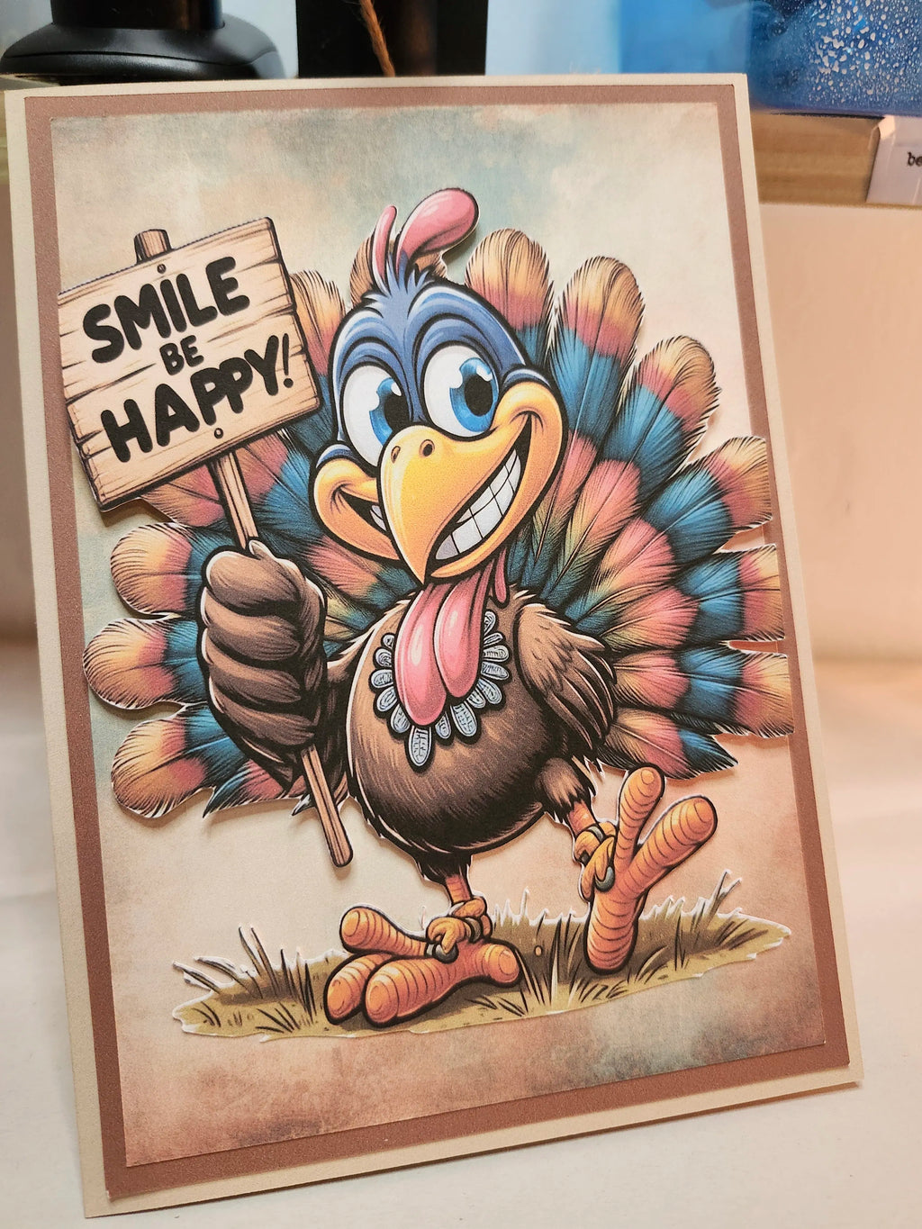 A handmade Thanksgiving greeting card featuring a cartoon turkey with colorful feathers and a sign that reads 'SMILE BE HAPPY!'.