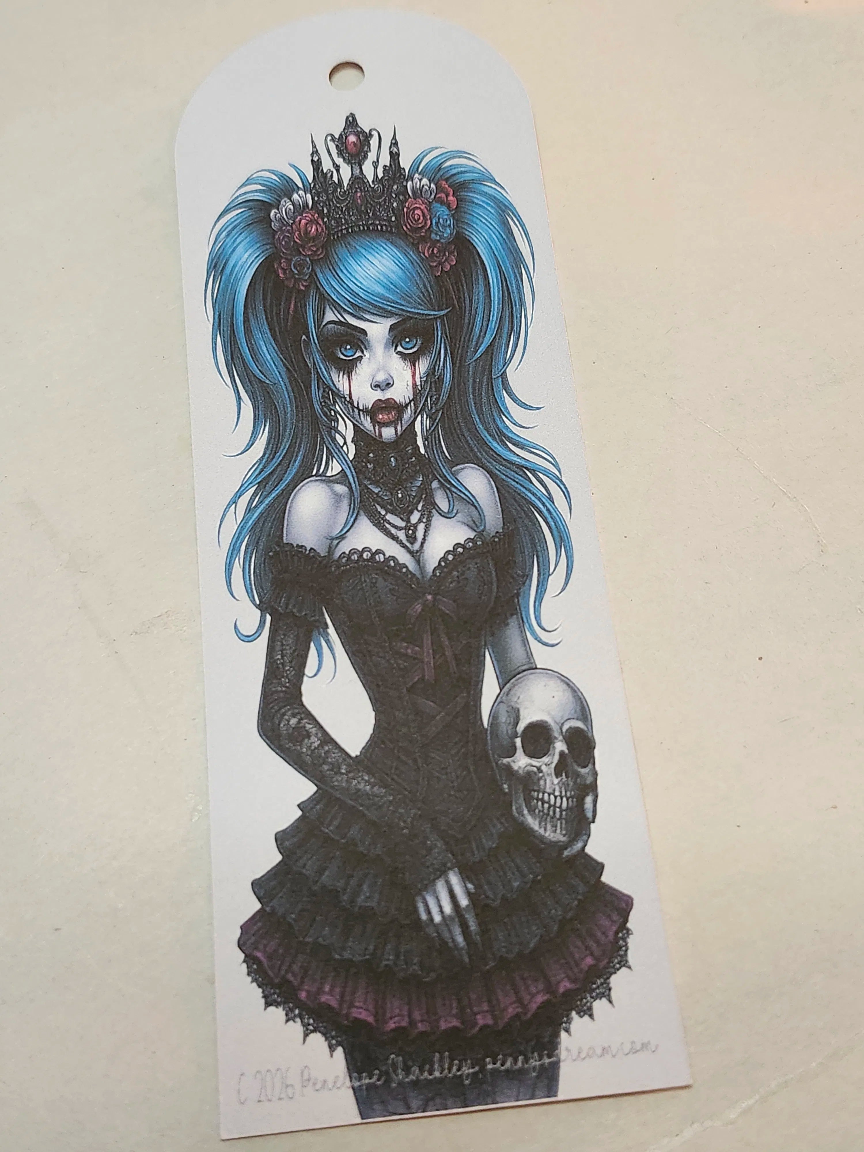 A white paper bookmark featuring a detailed illustration of a gothic zombie girl with bright blue hair, a black lace dress, and a skull, photographed on a beige surface.