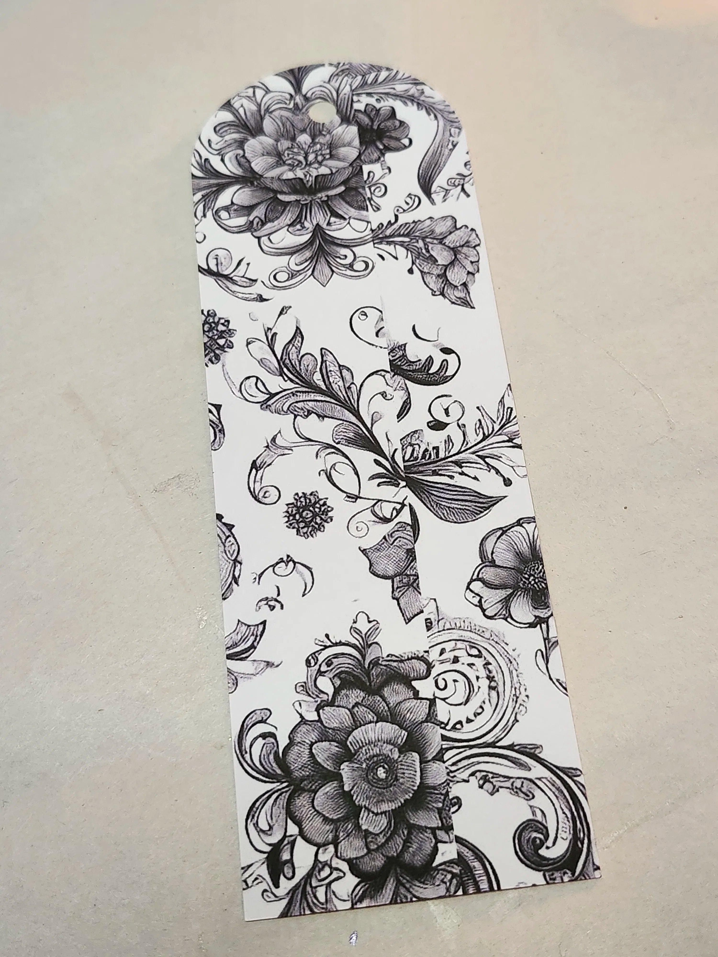 A white paper bookmark featuring a black floral and scrollwork pattern, photographed on a light beige textured surface.