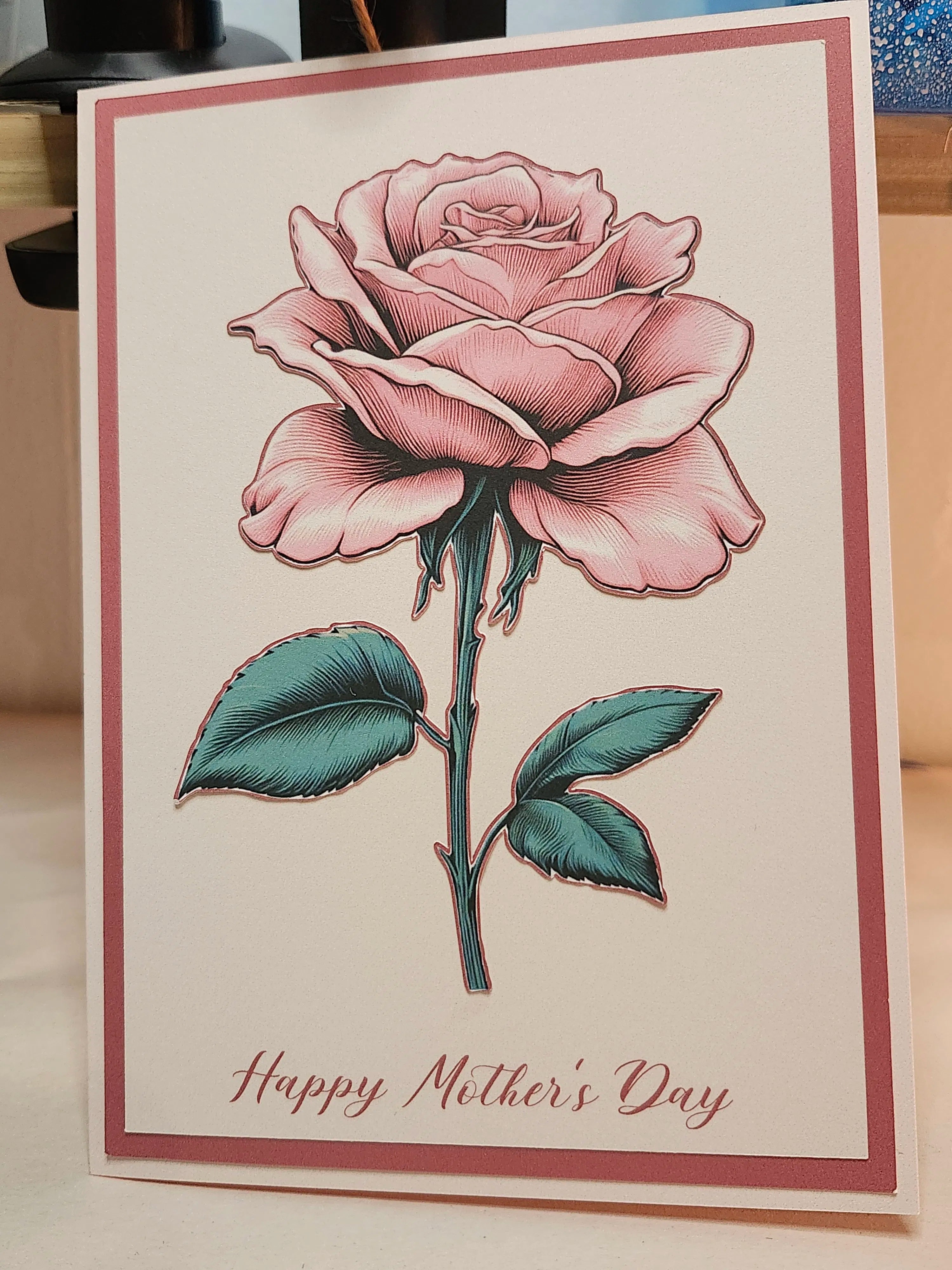 A handmade greeting card featuring a detailed pink rose with green leaves and the text 'Happy Mother's Day' in pink script at the bottom, set against a warm beige background.