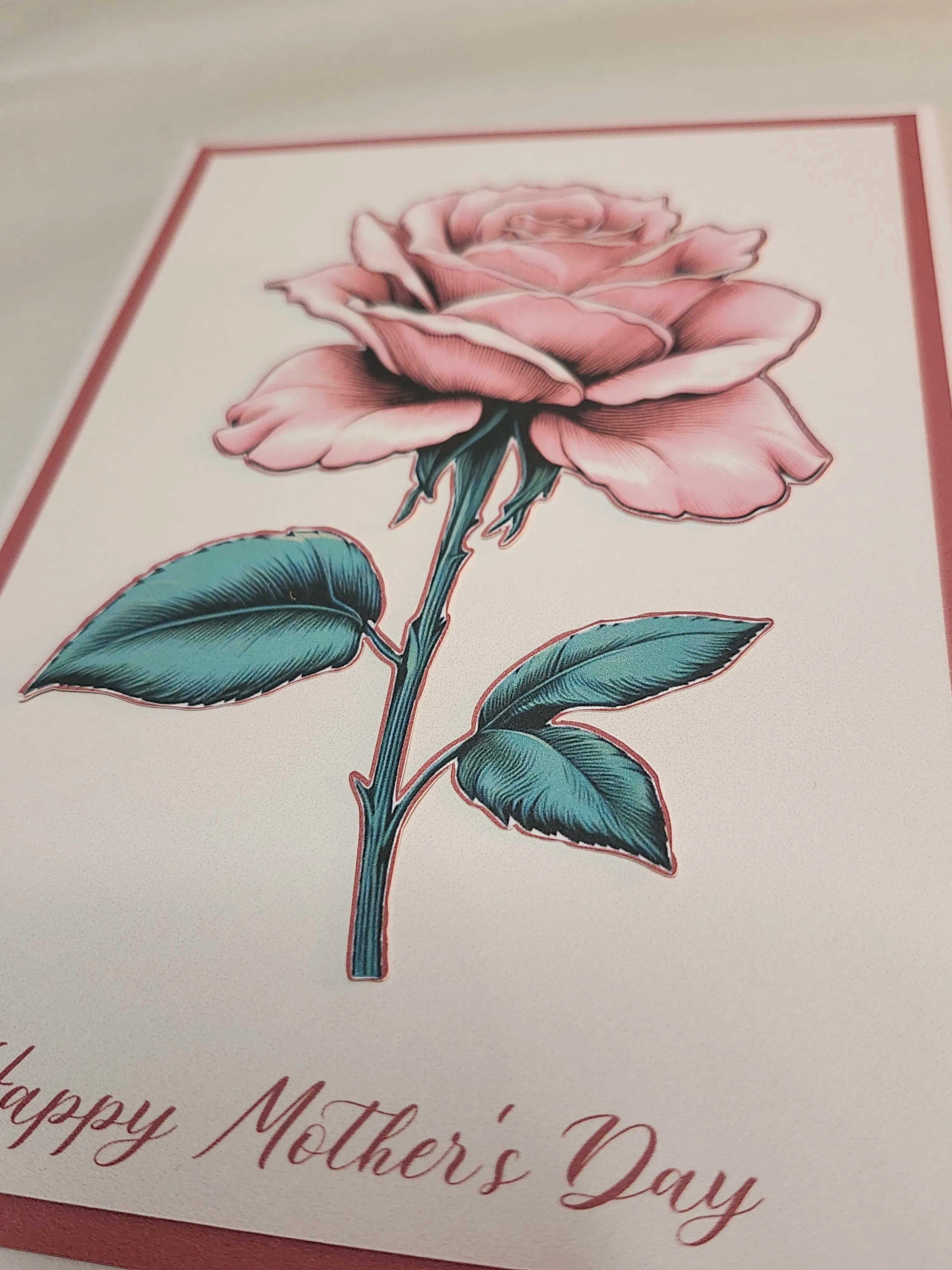 A close-up of a Mother's Day greeting card featuring a detailed pink rose with teal-green leaves on a white background, with 'Happy Mother's Day' written in cursive at the bottom.