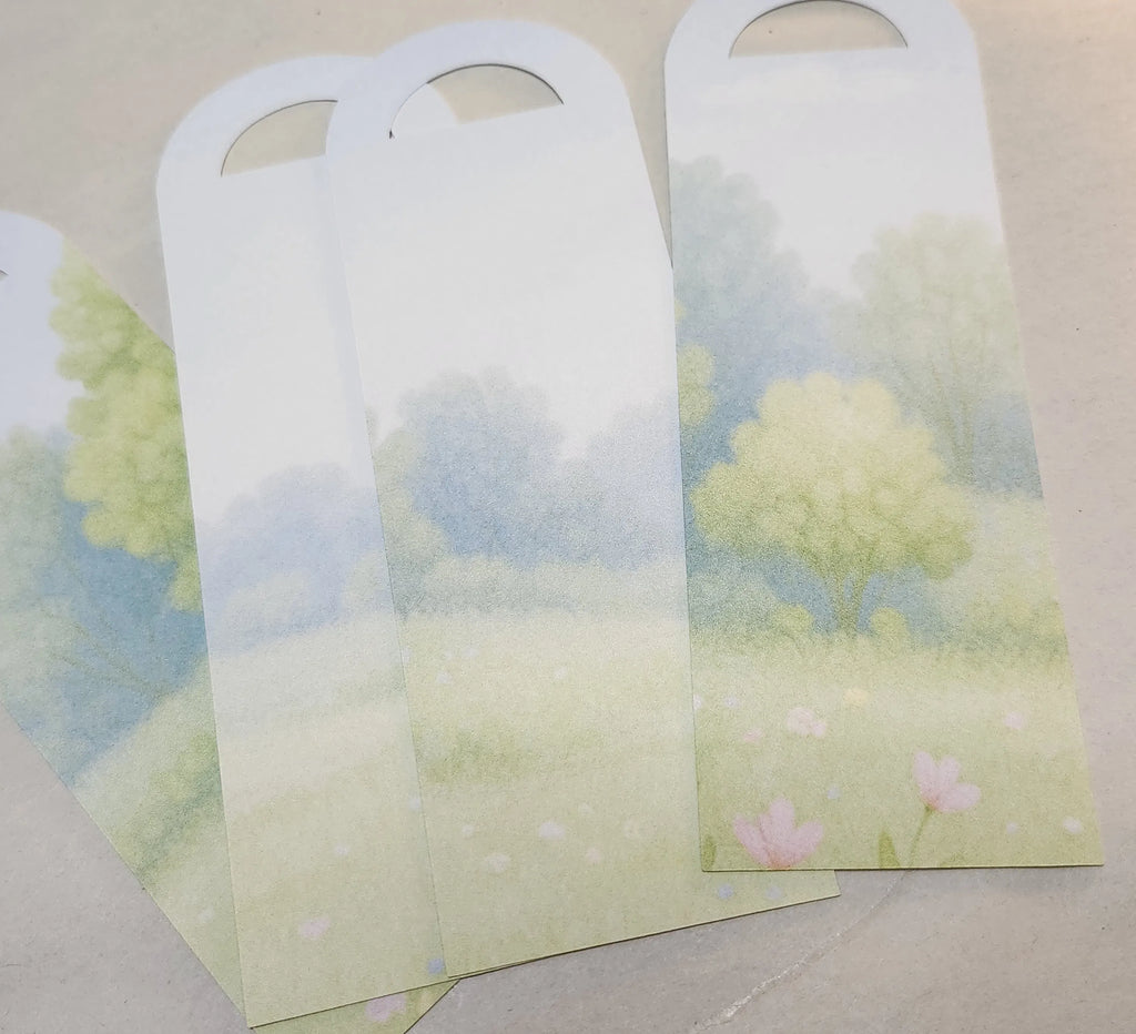 Bookmarks with rounded top corners and hanging holes, printed with a soft pastel landscape of trees and flowers in green, blue, and pink tones, arranged on a light beige surface.