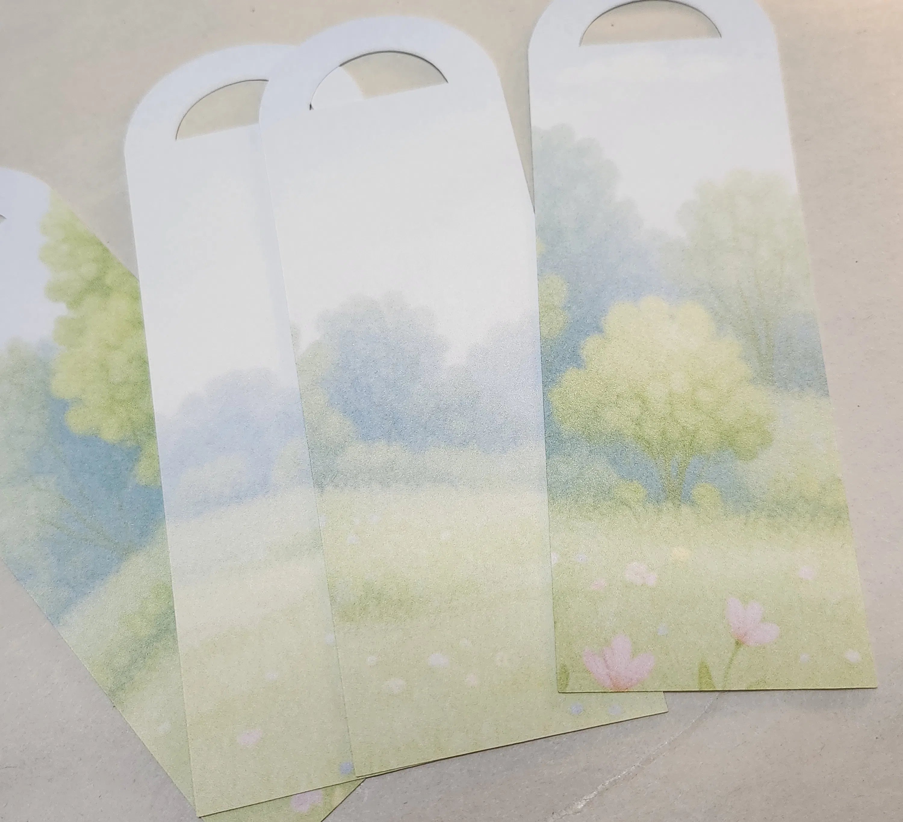 Bookmarks with rounded top corners and hanging holes, printed with a soft pastel landscape of trees and flowers in green, blue, and pink tones, arranged on a light beige surface.
