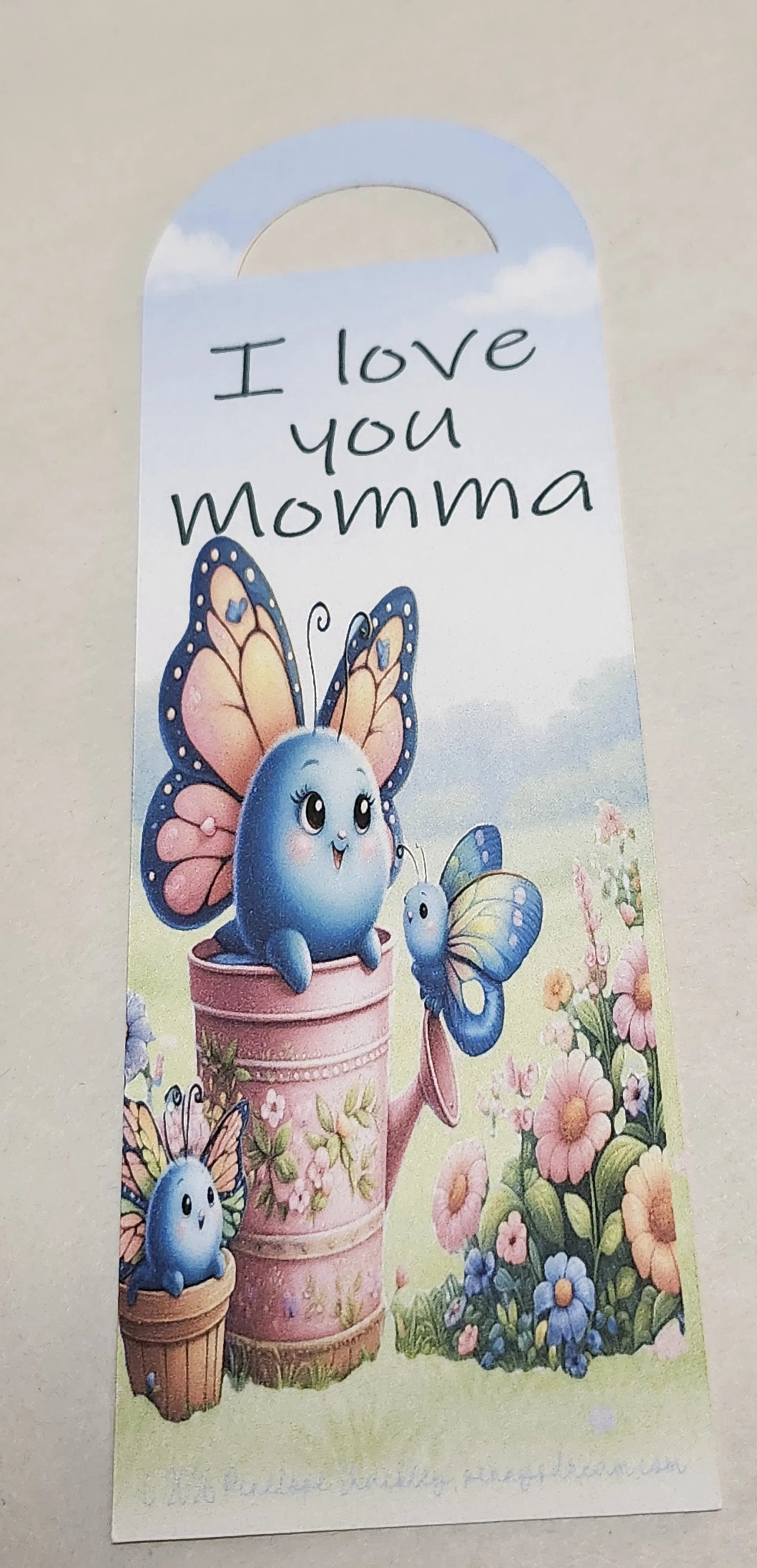 A personalized garden-themed bookmark with the text 'I love you Momma' in black script. The hanger features a cute blue cartoon bird character with a butterfly on a pink flower pot, surrounded by colorful flowers and green grass at the bottom, set against a light blue sky background.