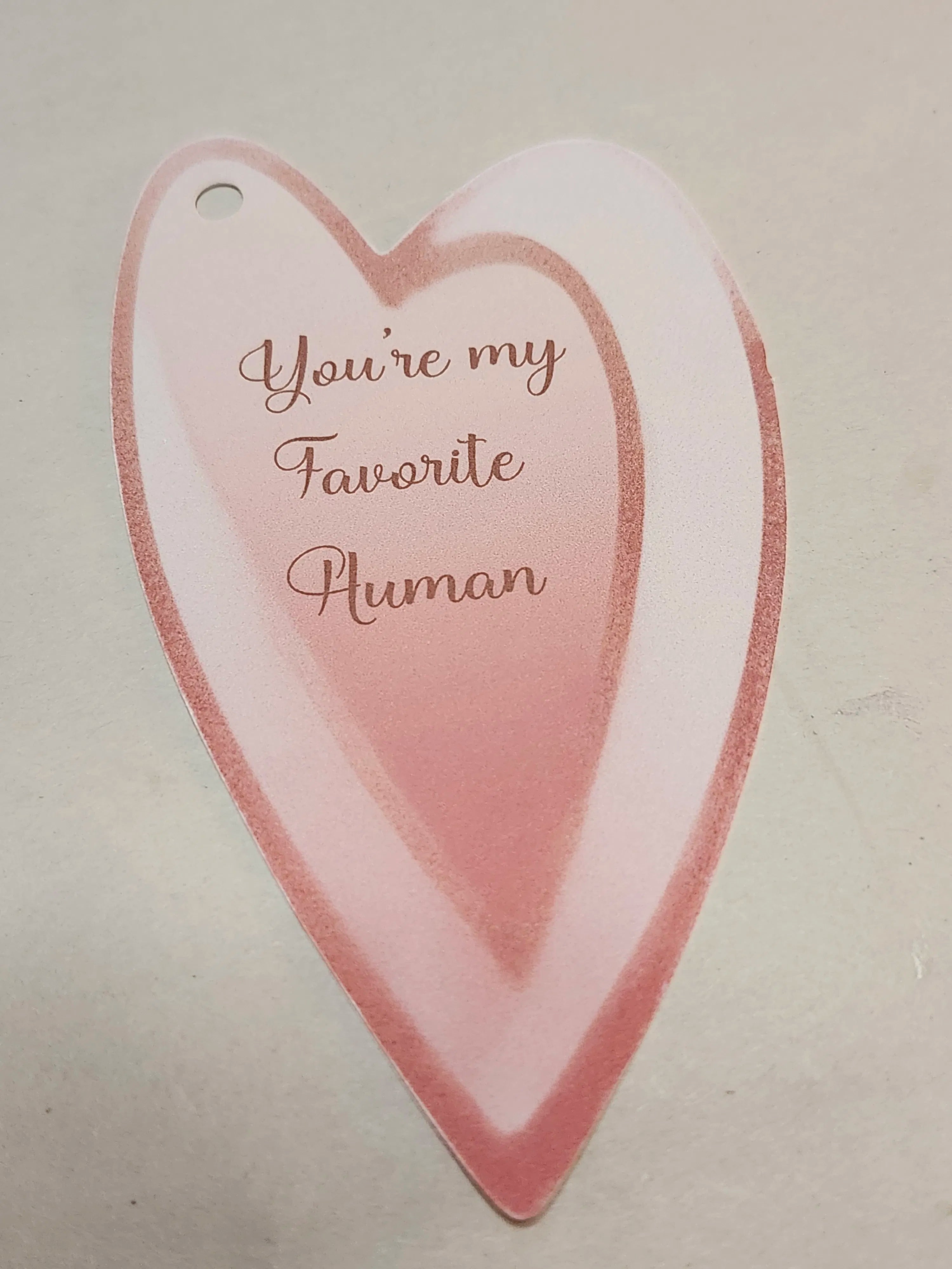A heart-shaped paper tag with a small hanging hole, featuring the phrase 'You're my Favorite Human' in rose-gold cursive text over a soft pink and white gradient background.