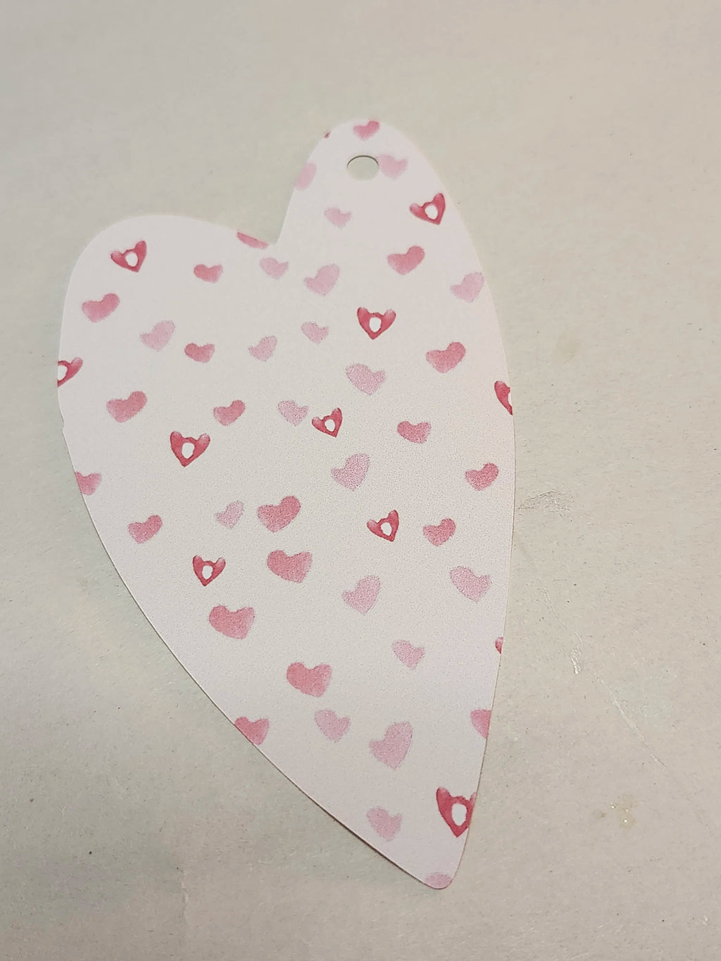 A heart-shaped paper tag or label with a small hole at the top, printed with a pattern of small pink and red hearts on a white background, placed on a light gray surface.