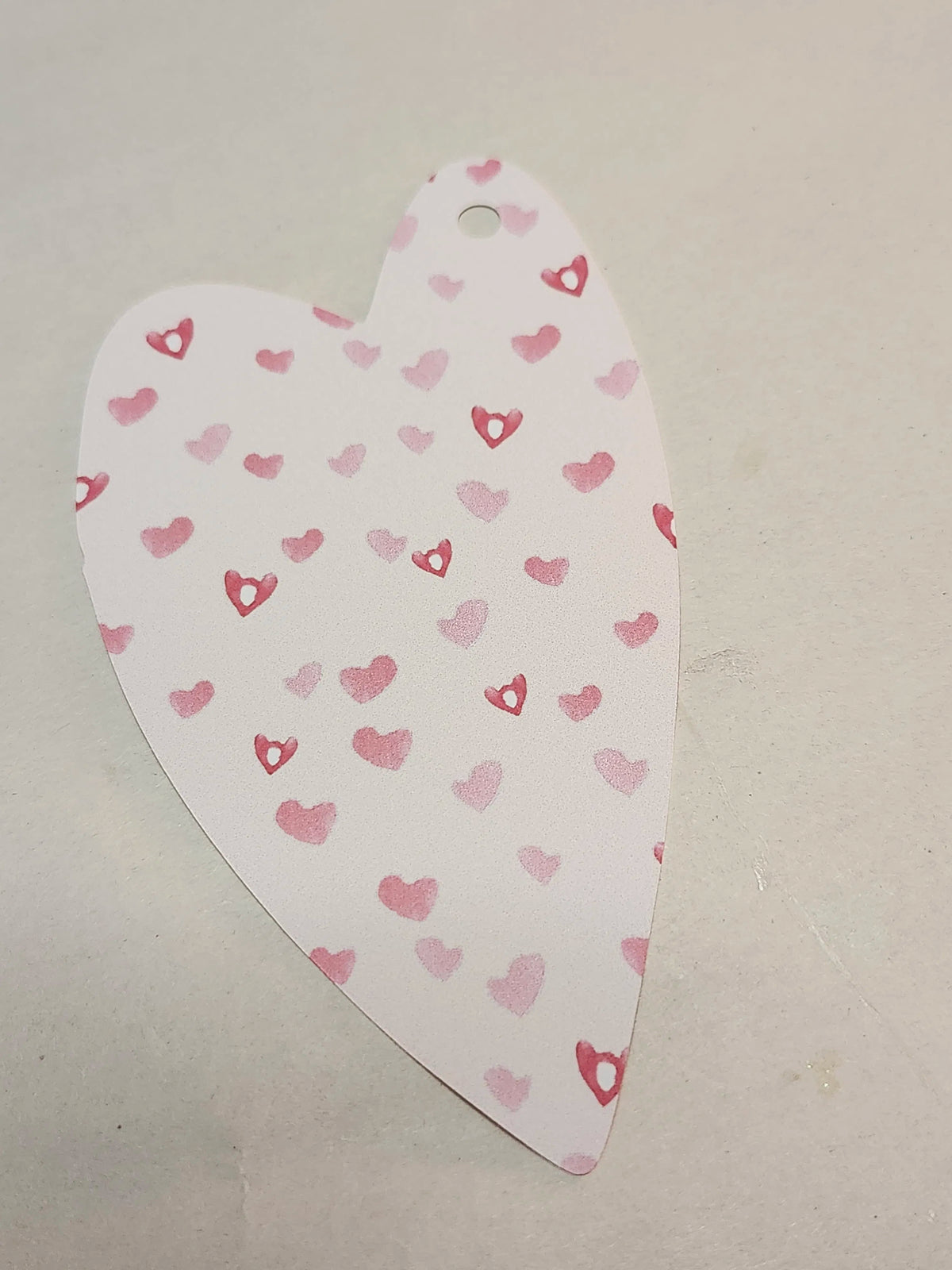 A heart-shaped paper tag or label with a small hole at the top, printed with a pattern of small pink and red hearts on a white background, placed on a light gray surface.