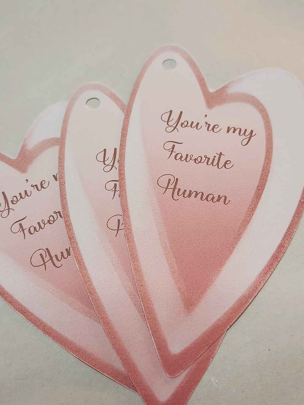 Three overlapping heart-shaped tags in soft pink and white, each printed with the phrase 'You're my Favorite Human' in cursive script.