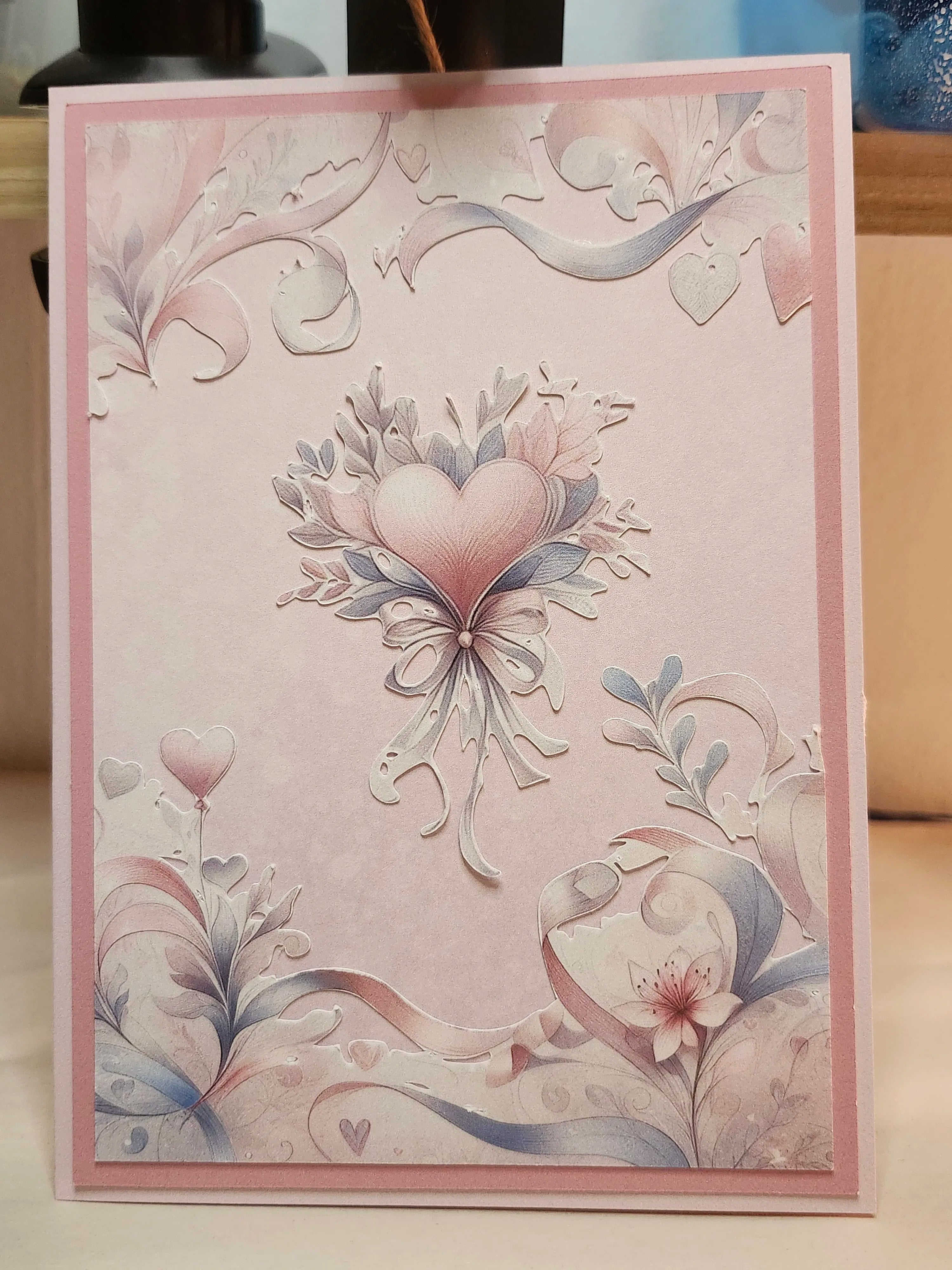 A square greeting card with a pink border and embossed floral heart design in pastel pink, white, and blue-gray tones, photographed standing upright on a tabletop.