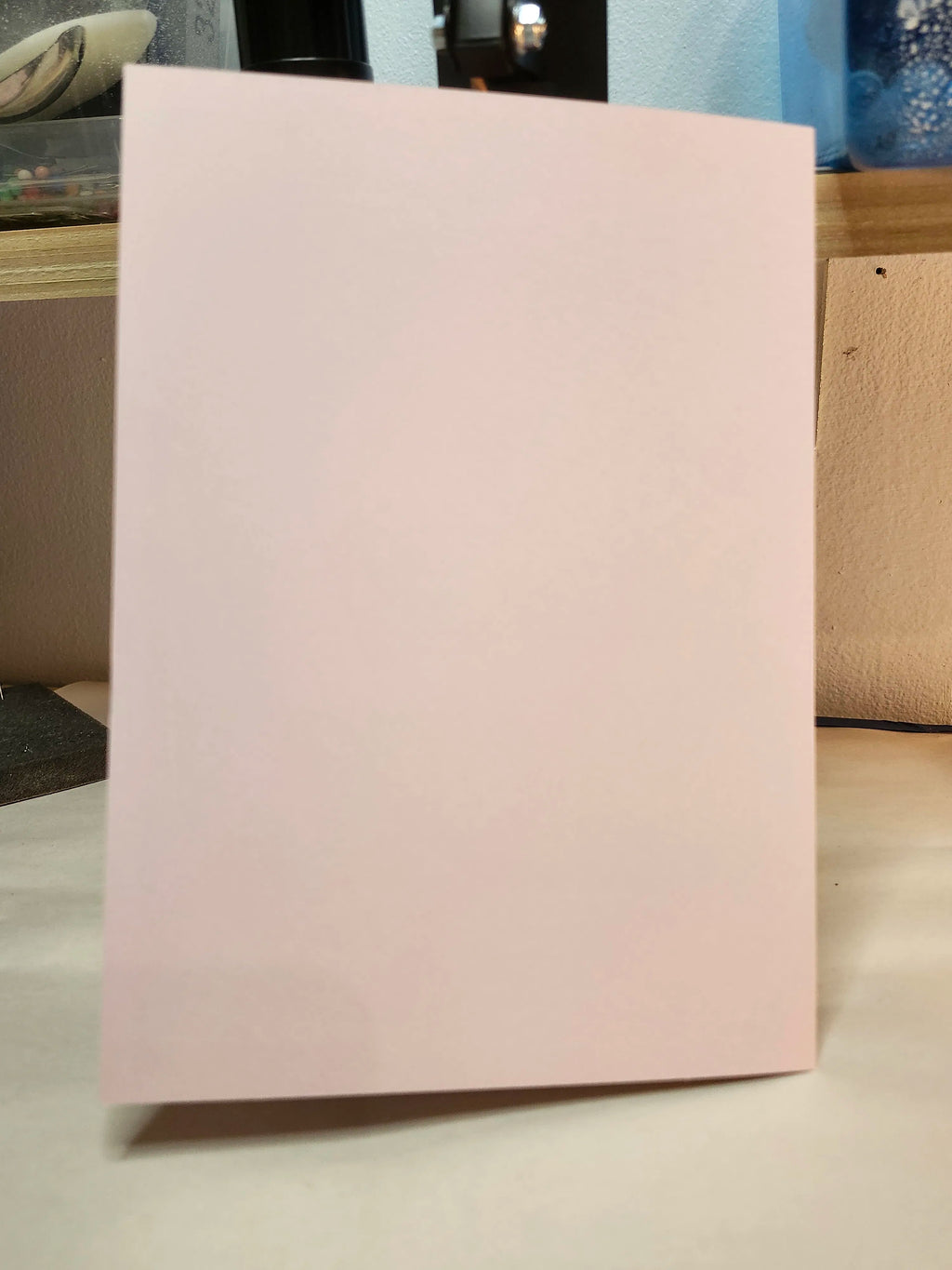 A plain light pink rectangular card or sheet standing upright on a tabletop, with a blurred indoor background.