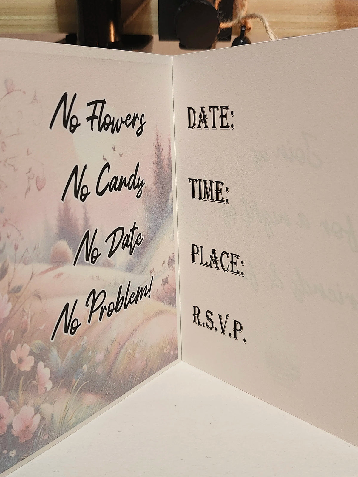 An open white wedding invitation card with a pastel floral landscape printed on the left panel and black text reading 'No Flowers No Candy No Date No Problem!' alongside fields for date, time, place, and RSVP.