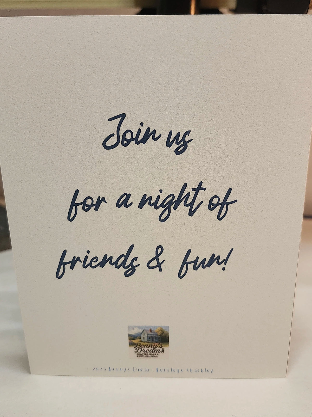 A white rectangular card or invitation standing upright on a light surface, with dark navy script text reading 'Join us for a night of friends & fun!' and a small printed logo near the bottom.