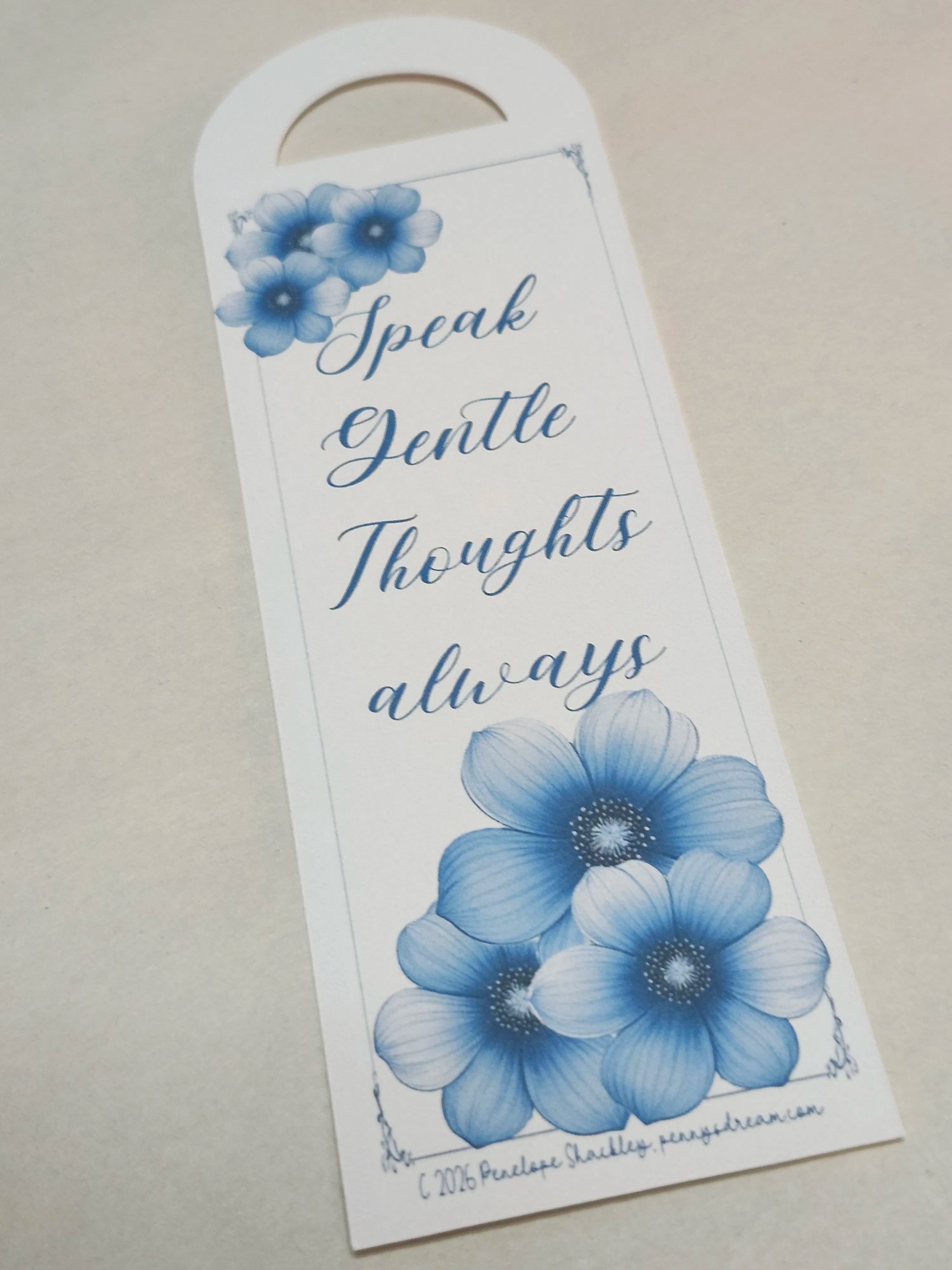A white paper door hanger with a blue floral design and the phrase 'Speak Gentle Thoughts always' in blue script, photographed on a light beige surface.