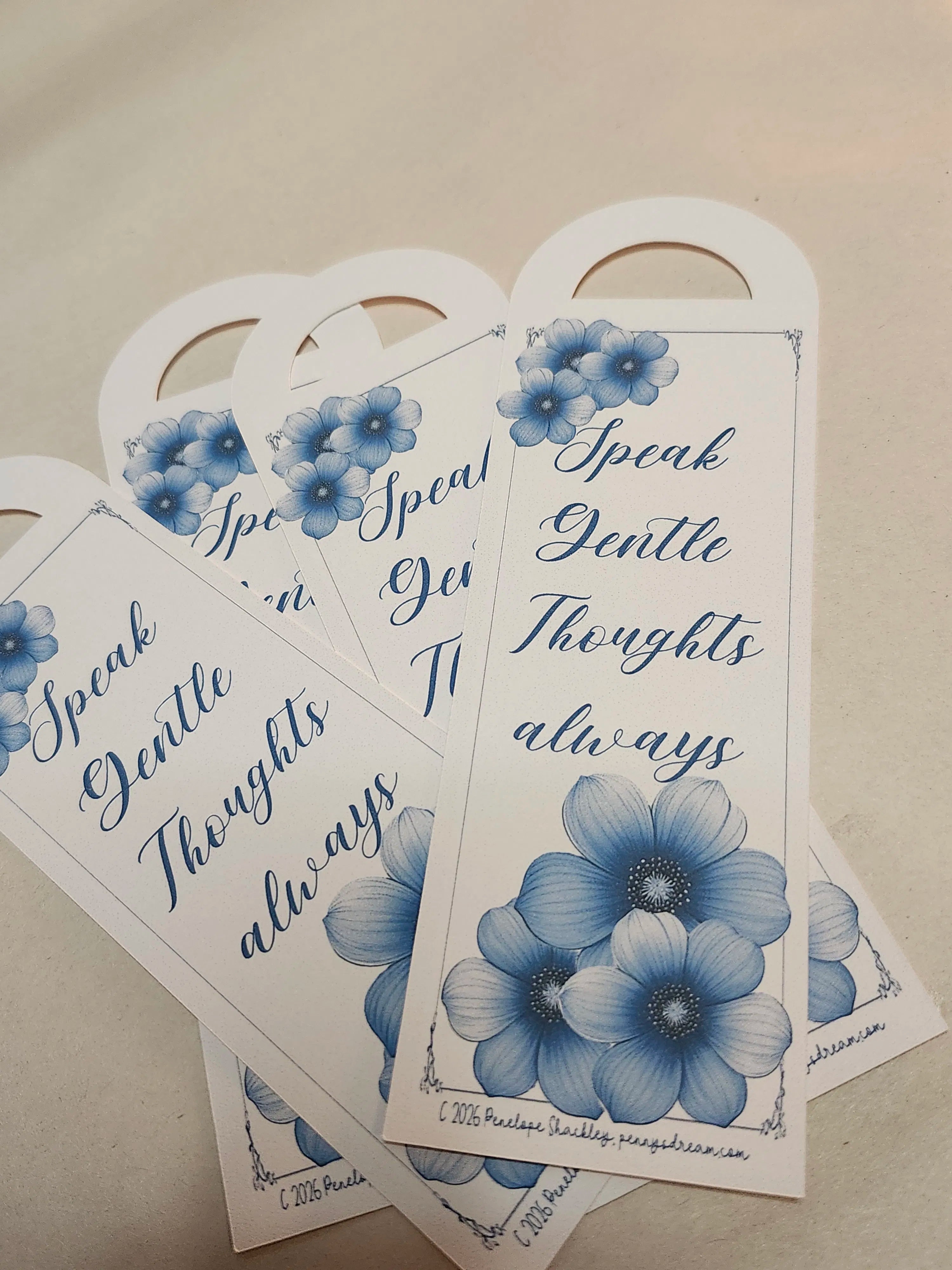 A set of white paper bookmarks or tags with blue floral illustrations and the phrase 'Speak Gentle Thoughts always' in blue script, arranged on a light beige surface.