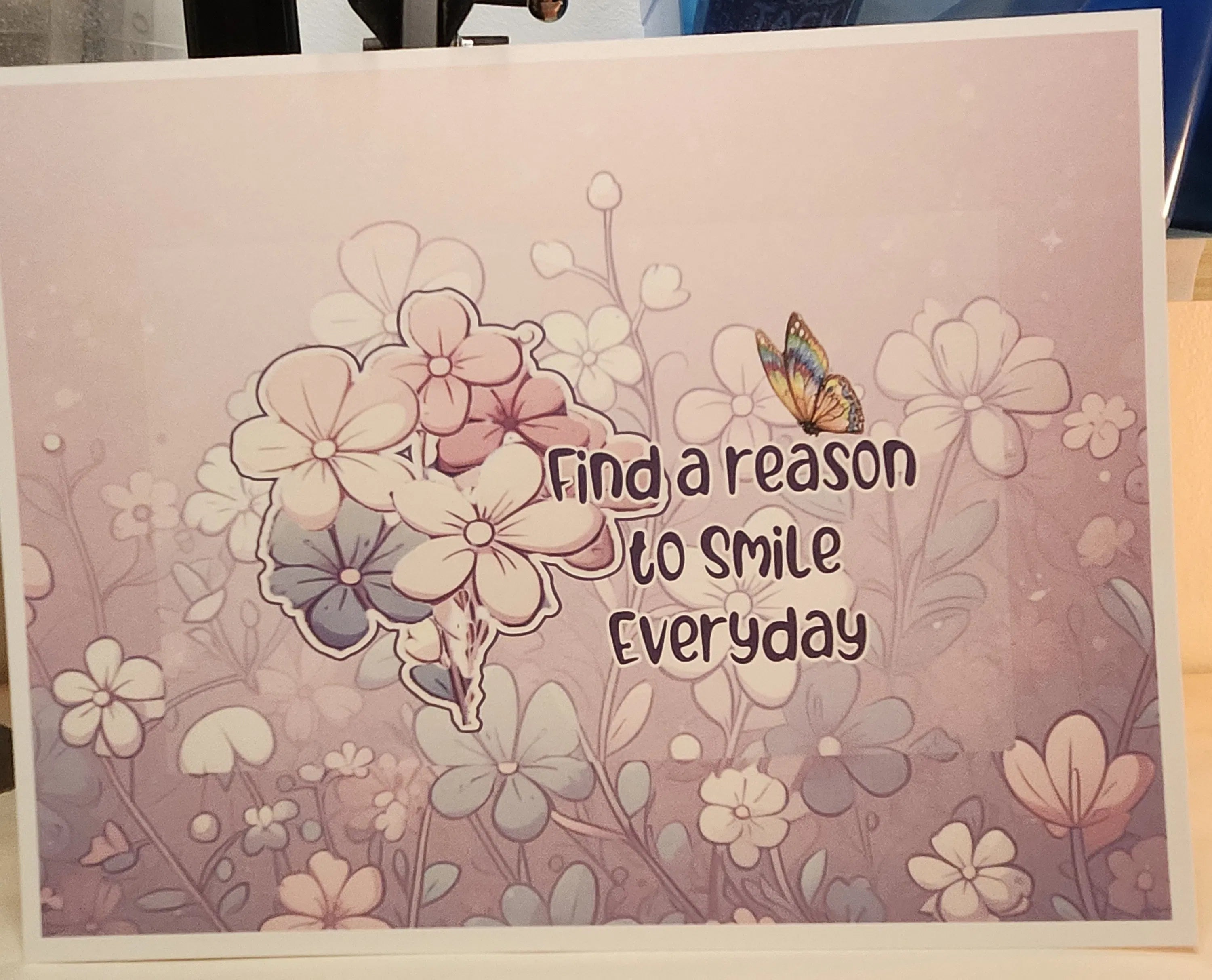 A square art print or card featuring the quote 'Find a reason to smile everyday' in dark brown lettering, surrounded by delicate floral illustrations in soft pink, lavender, and white tones, with a small butterfly on the right side.