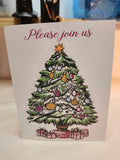 A square greeting card with the text 'Please join us' at the top, featuring an illustrated Christmas tree decorated with ornaments and gifts at the bottom, photographed standing upright on a tabletop.