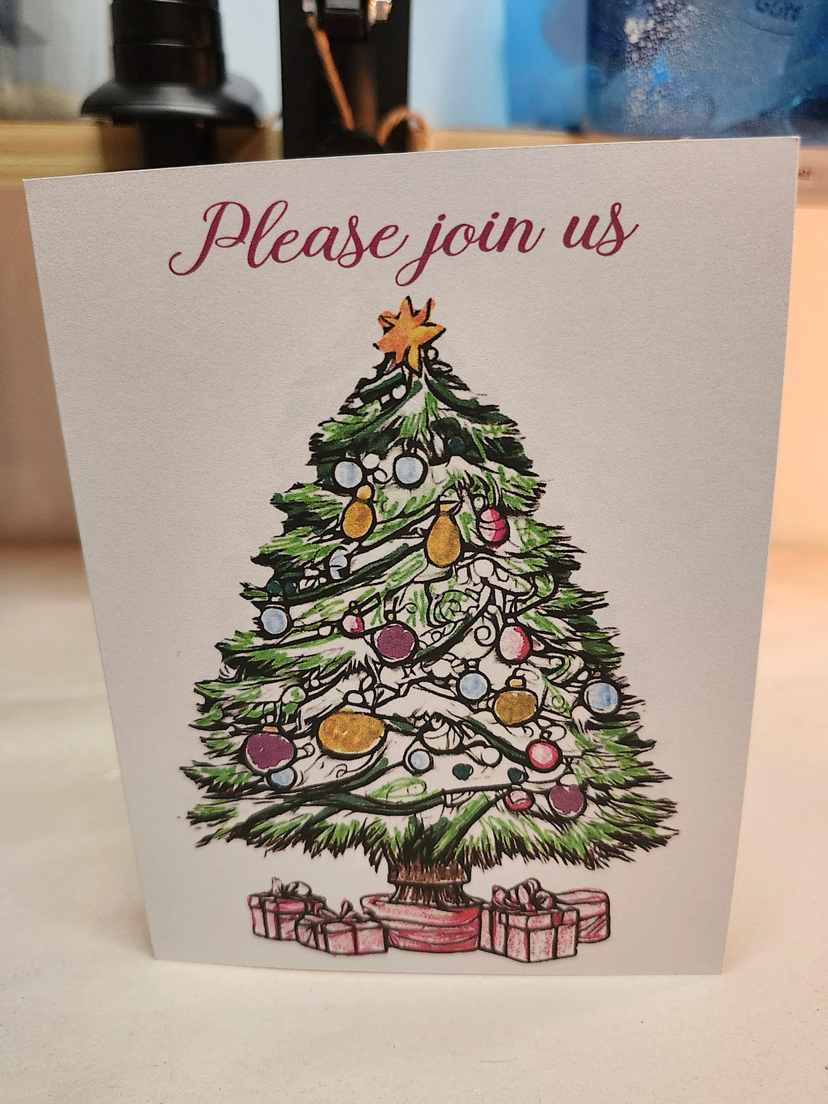 A square greeting card with the text 'Please join us' at the top, featuring an illustrated Christmas tree decorated with ornaments and gifts at the bottom, photographed standing upright on a tabletop.