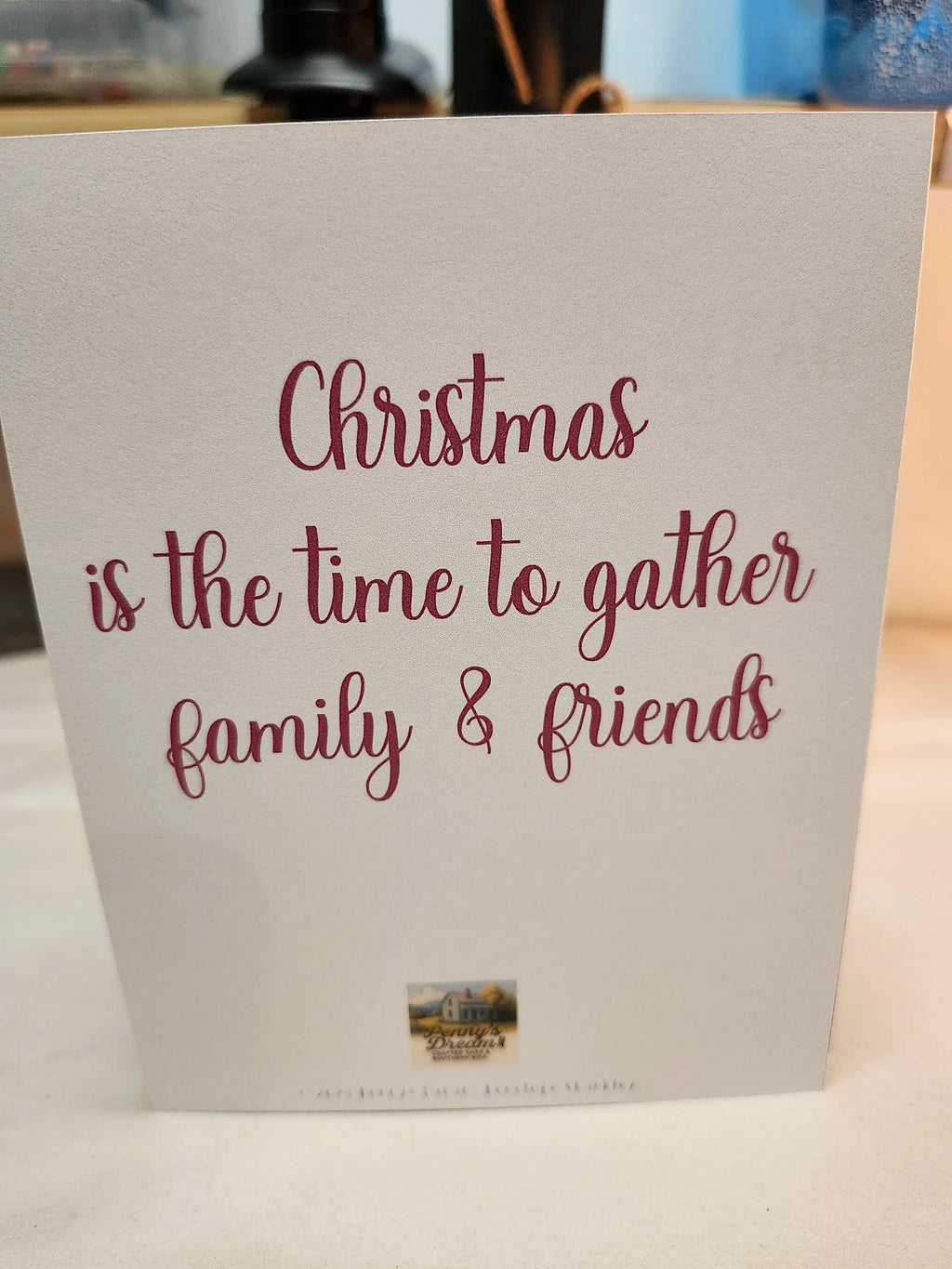 A white greeting card with the phrase 'Christmas is the time to gather family & friends' printed in dark red script, photographed standing upright on a light surface.