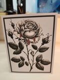A square art print or card featuring a black and white botanical illustration of a rose with red blood-like drips, photographed standing on a light surface with a blurred indoor background.
