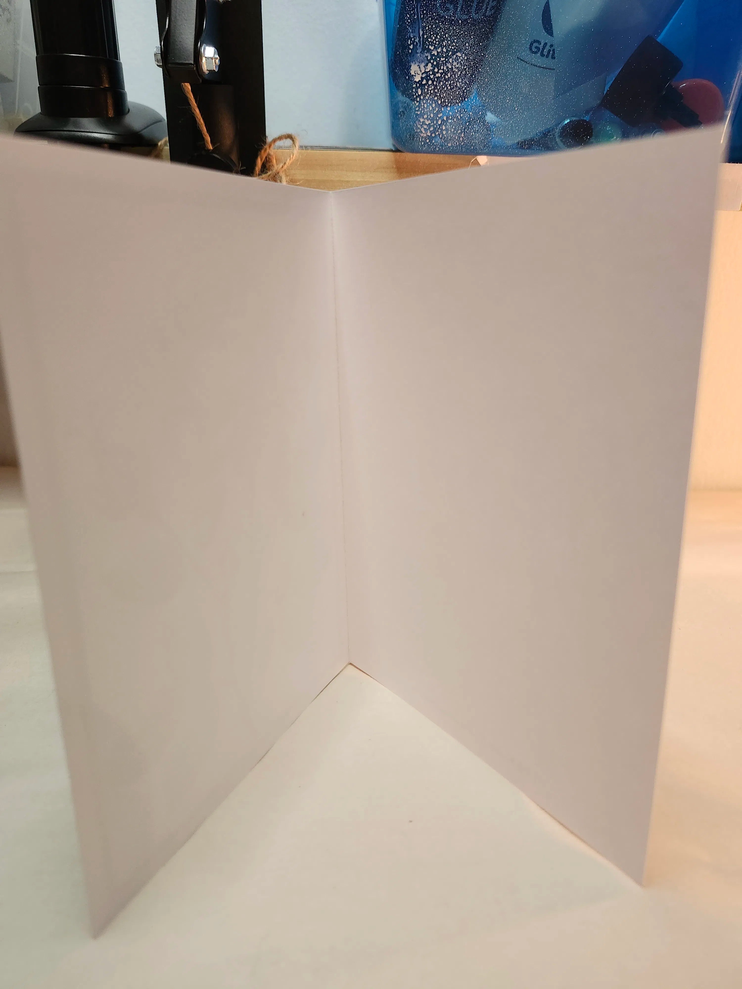 A white card or sheet of paper standing open to form a V-shape corner, photographed indoors on a light surface with blurred background objects.