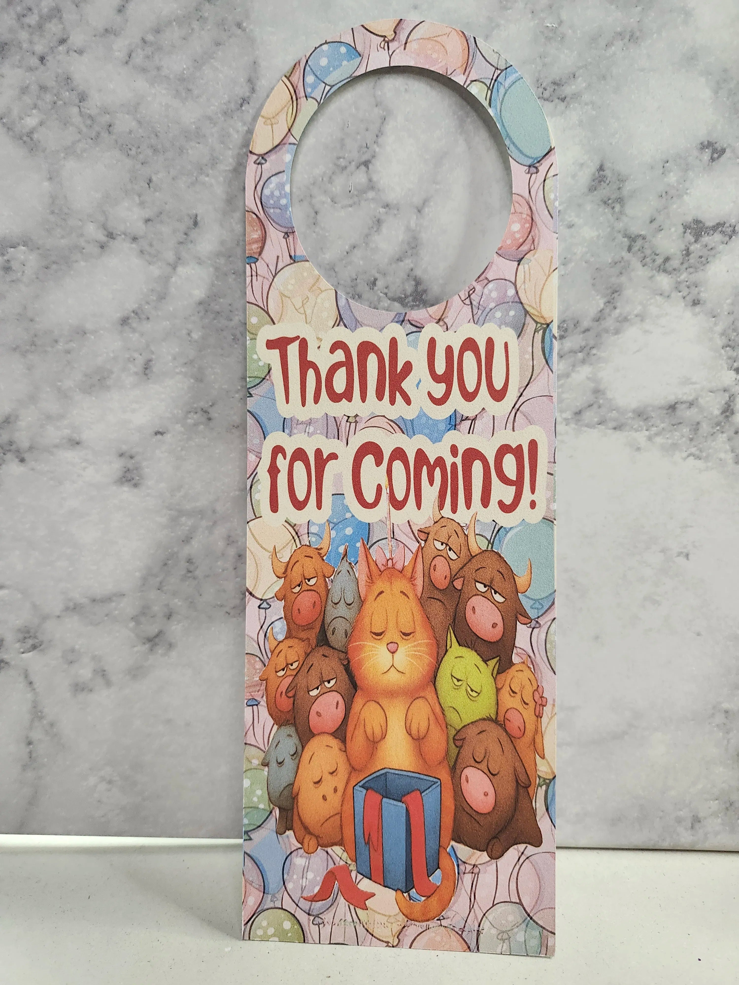 A novelty paper doormat with a hanger loop at the top, featuring the text 'Thank you for coming!' in red and an illustration of a ginger cat surrounded by colorful cartoon animals and balloons, set against a gray marble background.