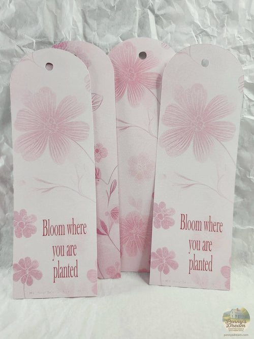 Three pink paper plant tags with floral designs and the printed phrase 'Bloom where you are planted' arranged on a white crinkled paper background.
